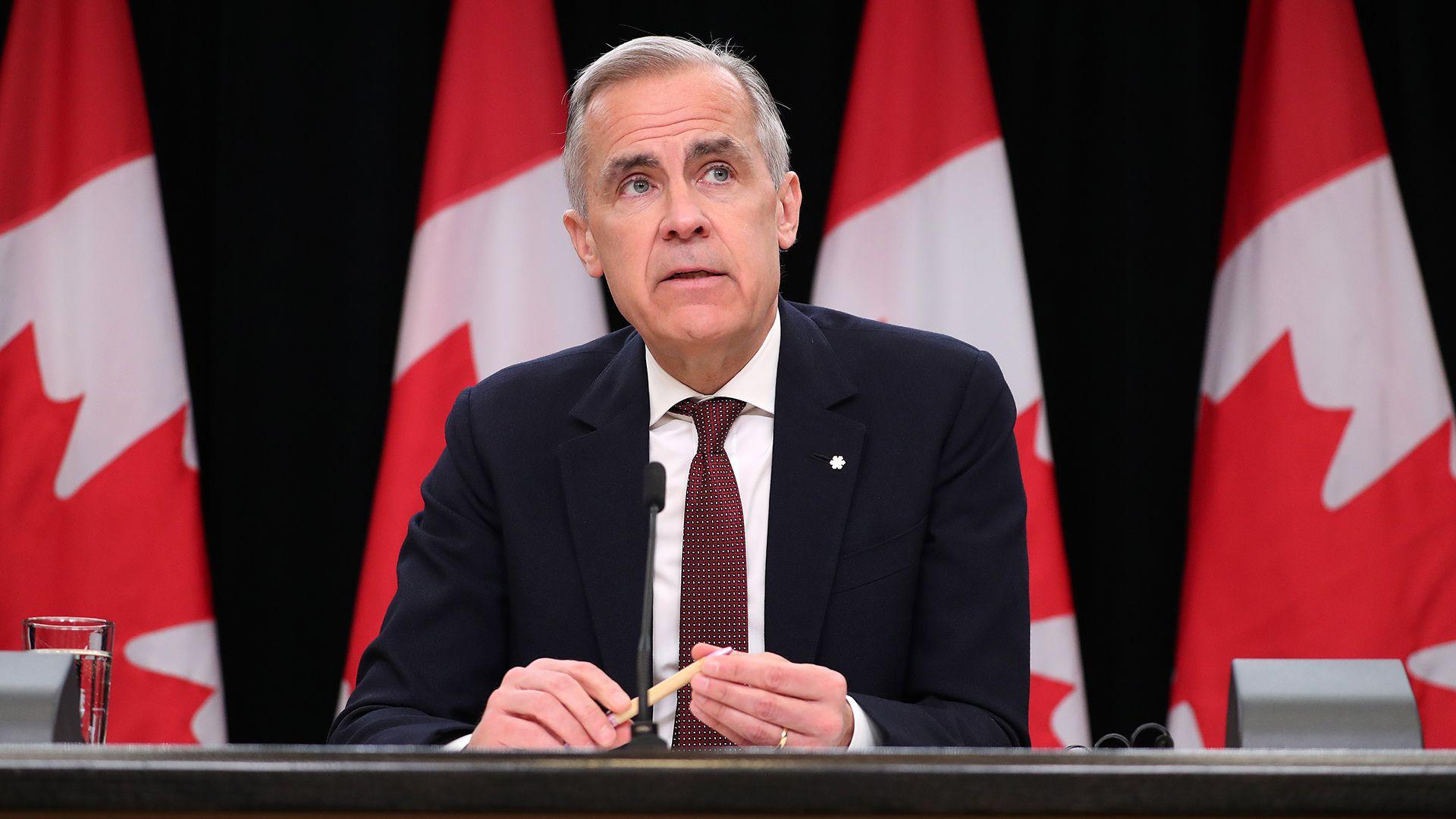 Canada's PM Mark Carney looks to the right of frame while sitting at a press conference briefing table in front of four Canadian flags in Ottowa in May. He wears a black suit and red tie, and holds a pen in his hands with a glass of water beside him on the table.