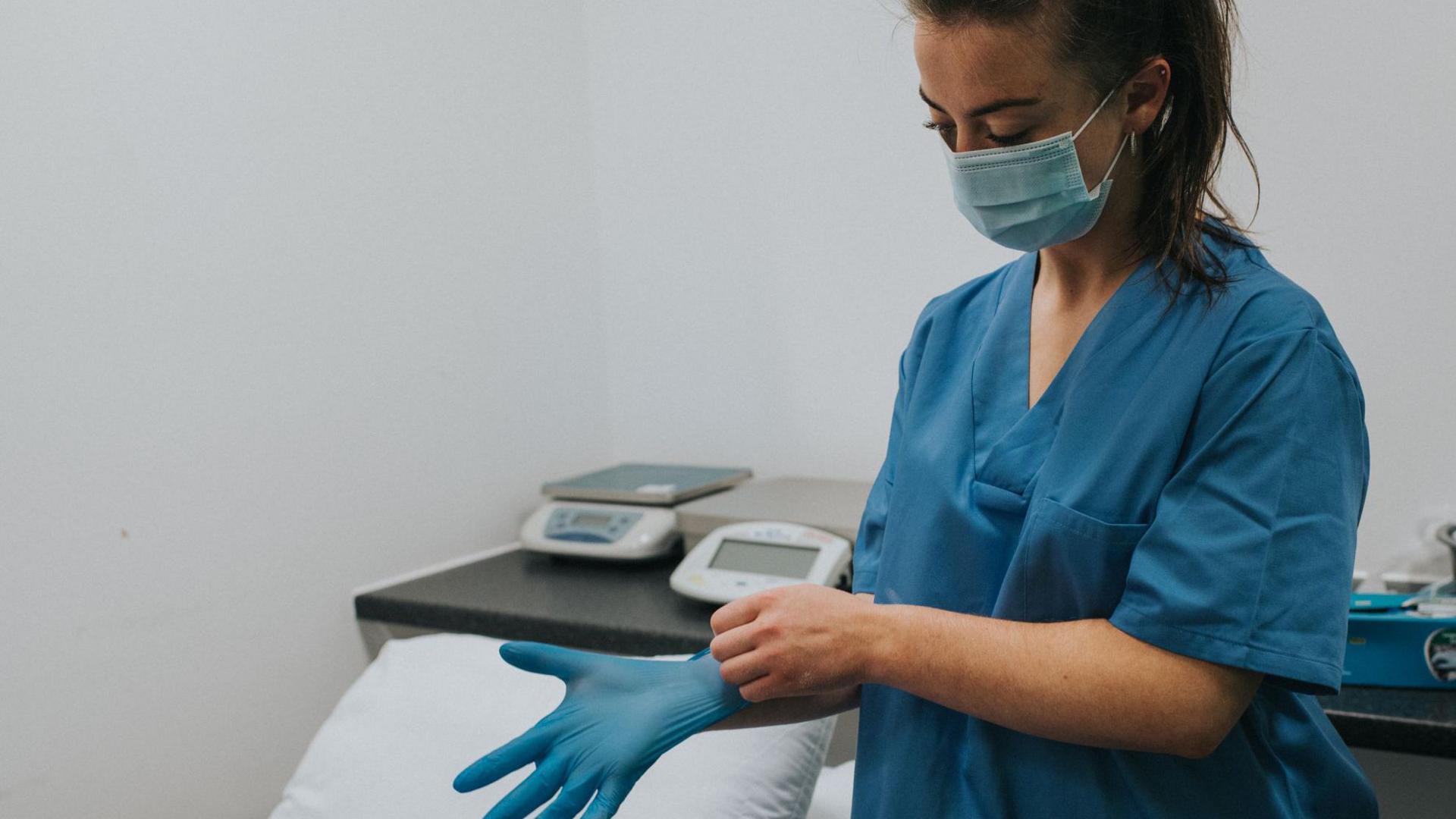 A nurse, wearing blue scrubs, standing beside a hospital bed. She has long brown hair, tied up. She is wearing a face mask and is putting a blue glove on one hand. 