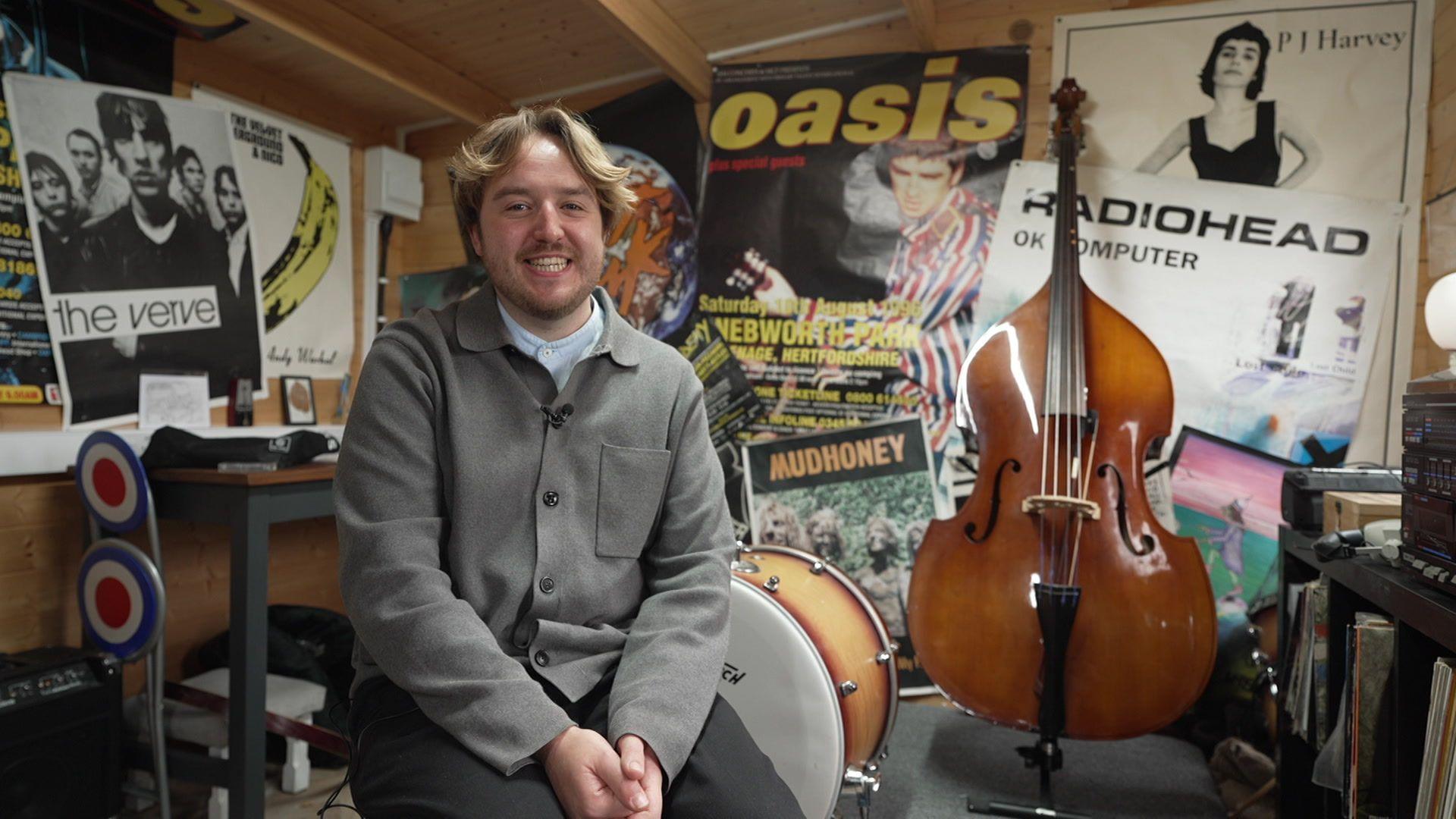Sitting on a chair in his music studio, Dan Astles is smiling for the camera. There are music posters on the wall for The Verve, Oasis, Radiohead, Mudhoney and P J Harvey. There is a cabinet on the right with a collection of vinyls and various musical instruments and recording equipment dotted around the room.
