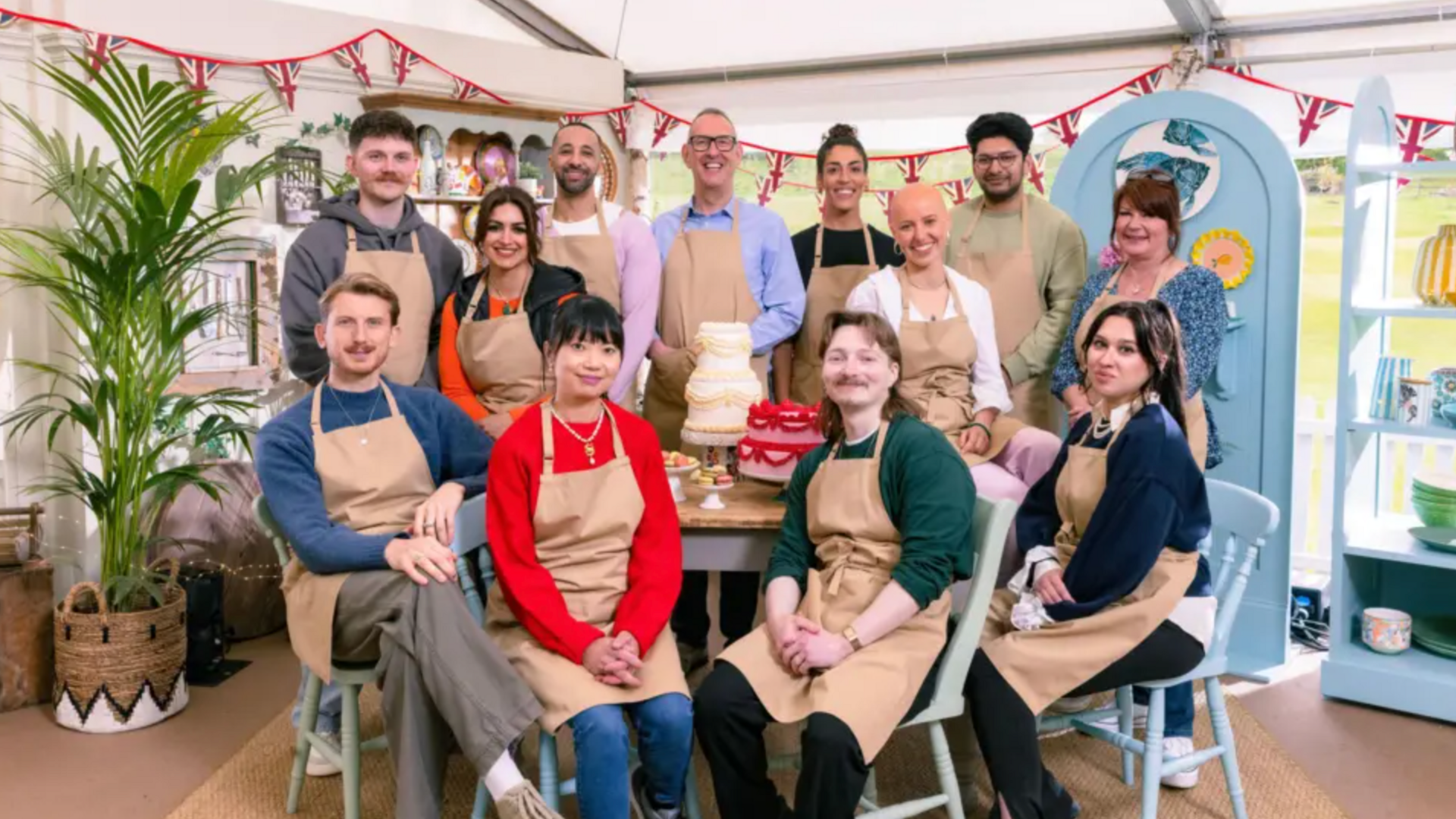 A group of 12 men and women, with four sat in front of a table and the other 8 standing. They are all wearing beige aprons and posing for the picture being taken. The background has a plant in a basket, bunting with the Union Jack on it and a blue door on the right hand side. A light blue shelf can also be seen.