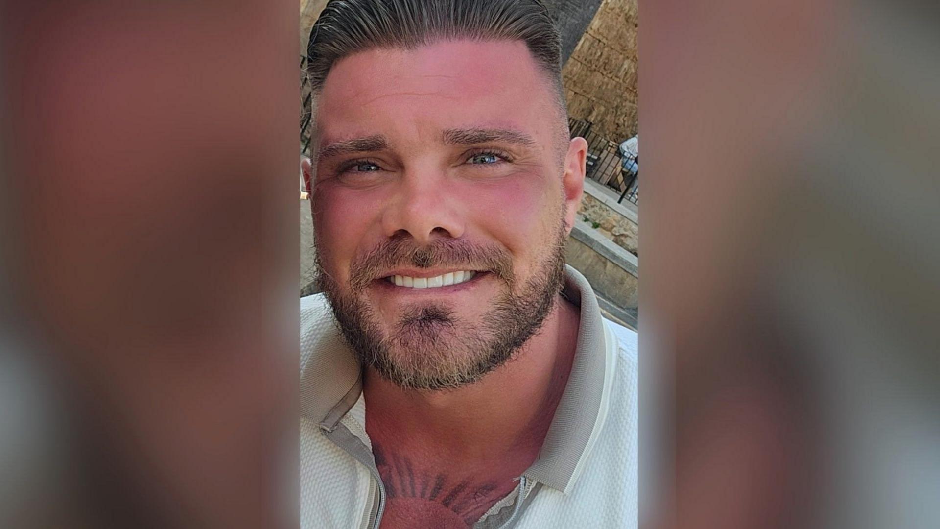 Philip McClelland samiling at the camera. He has short, dirty fair hair slicked back and a beard. He is wearing a white polo shirt with a zippable beige collar. He has a tattoo on his chest which is partially visible under his collar.