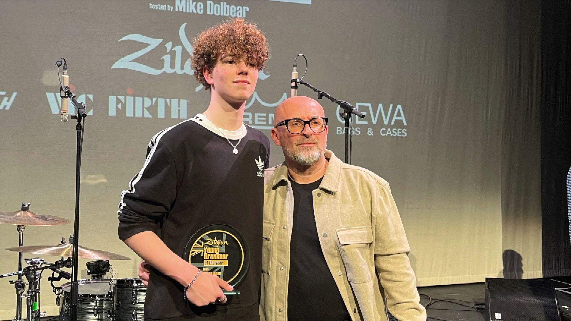 Jack holds his trophy and poses for a photo with the competition organiser. He is on a stage in front of a backdrop that features the names of sponsors and a drum kit. 