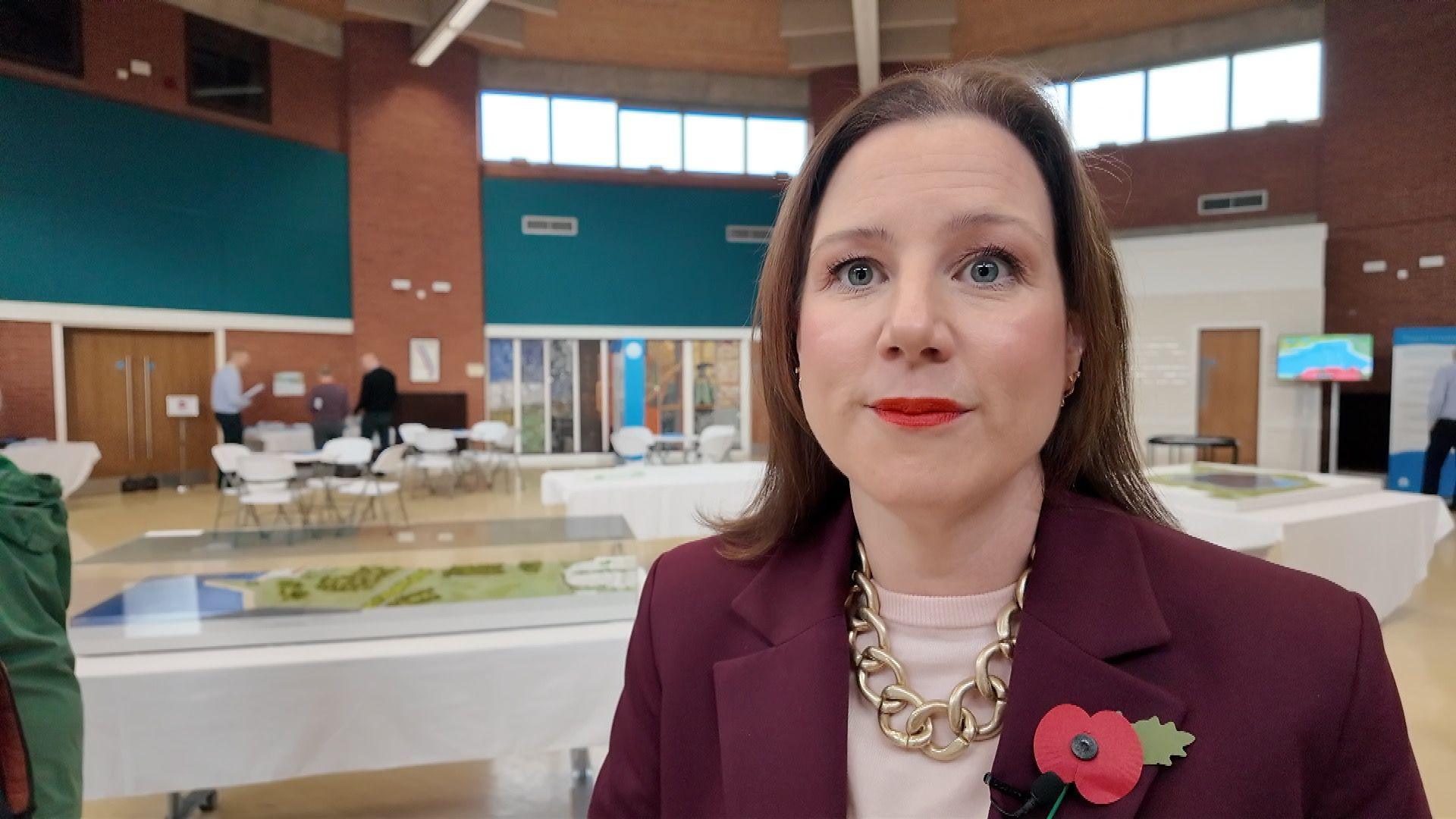 A woman with dark hair wearing a burgandy jacket and gold chain stands in front of a model of the reservoir