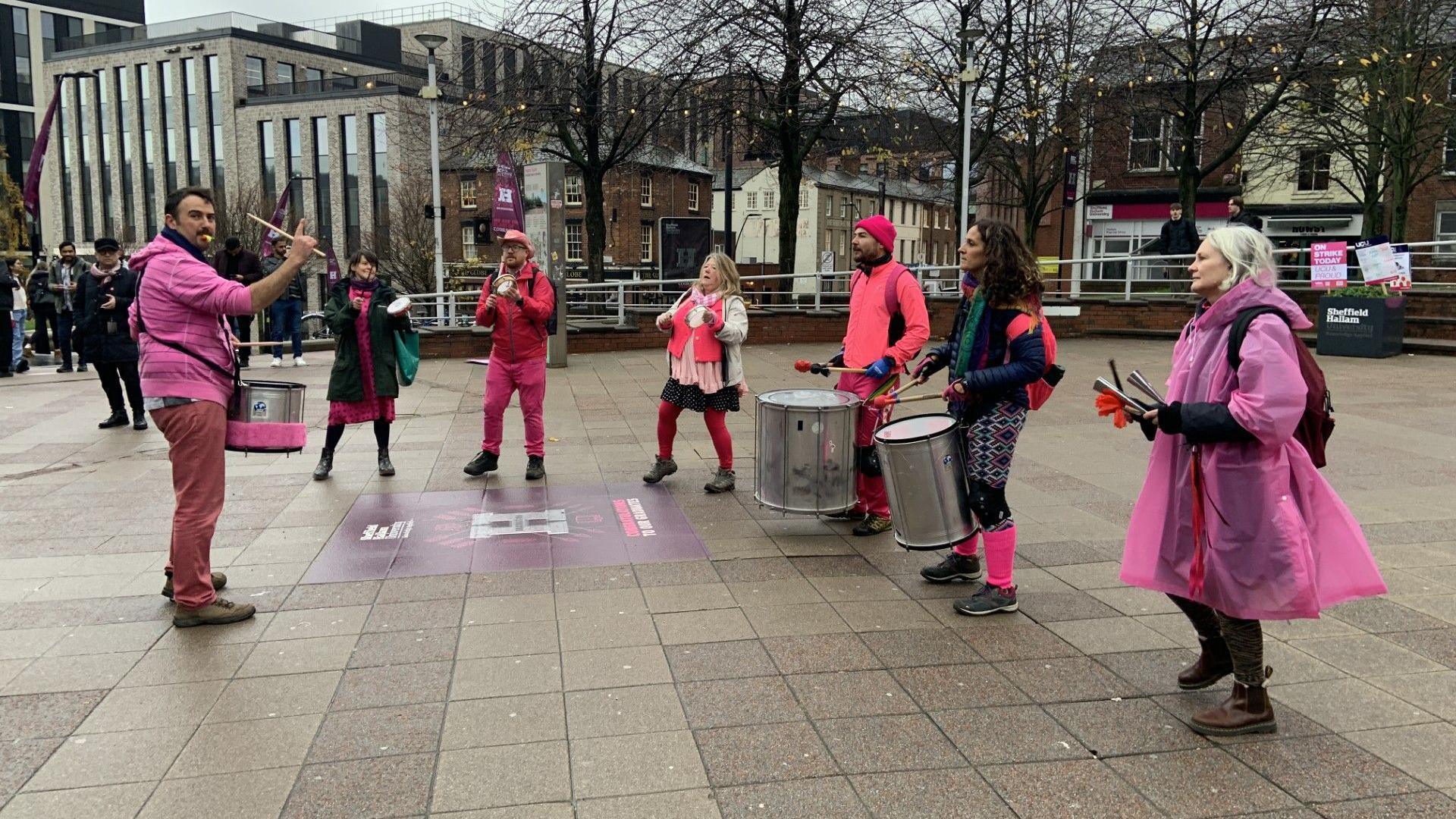 A group of people dressed in pink clothing are playing a variety of instruments on a picket line outside Sheffield Hallam University