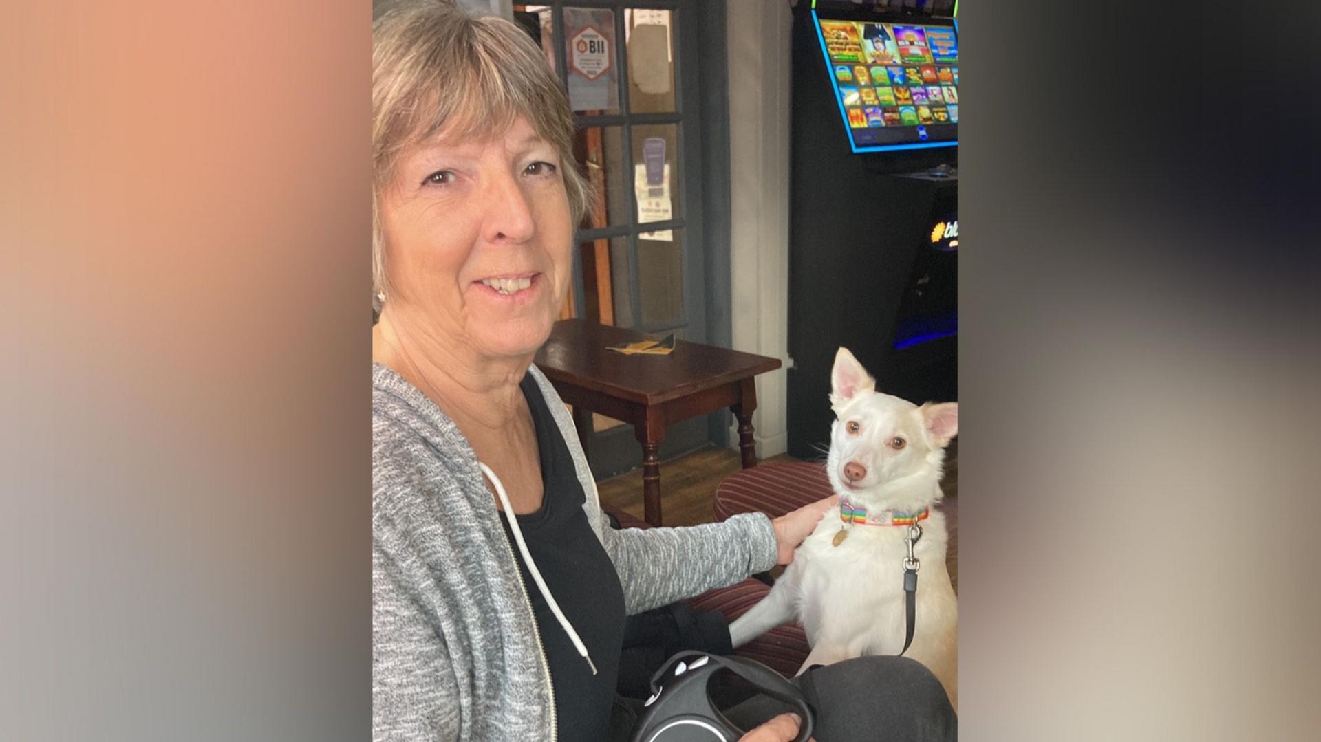 Andy Marriott's mother, Jan, with rescue dog Laika at home in a sitting room. She is wearing a black top and grey fleece and there is a table in the background.