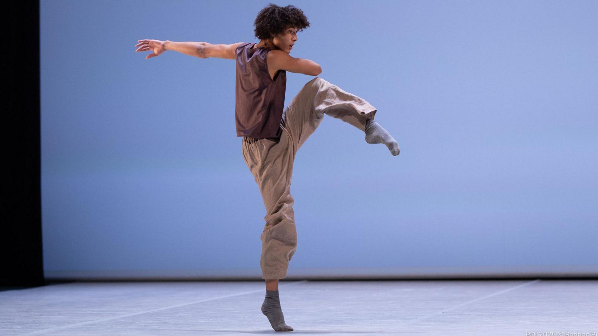 Jakob dancing, on a stage, with a blue background. He is wearing a brown top, brown trousers and grey socks. His left arm is outstretched and right arm across his body. His leg is raised. He has short curly dark hair. 