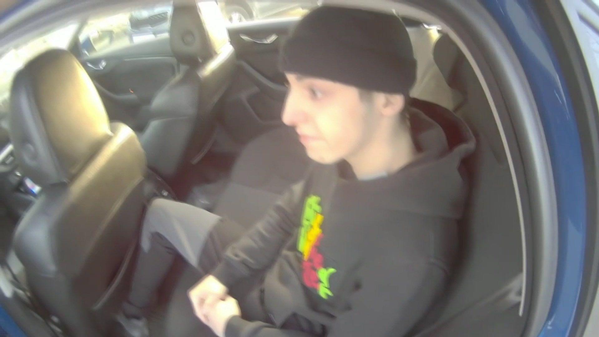 Police footage of Green sitting in the back of the police vehicle. He is wearing a black beanie, a black hoodie, and black tracksuit bottoms.