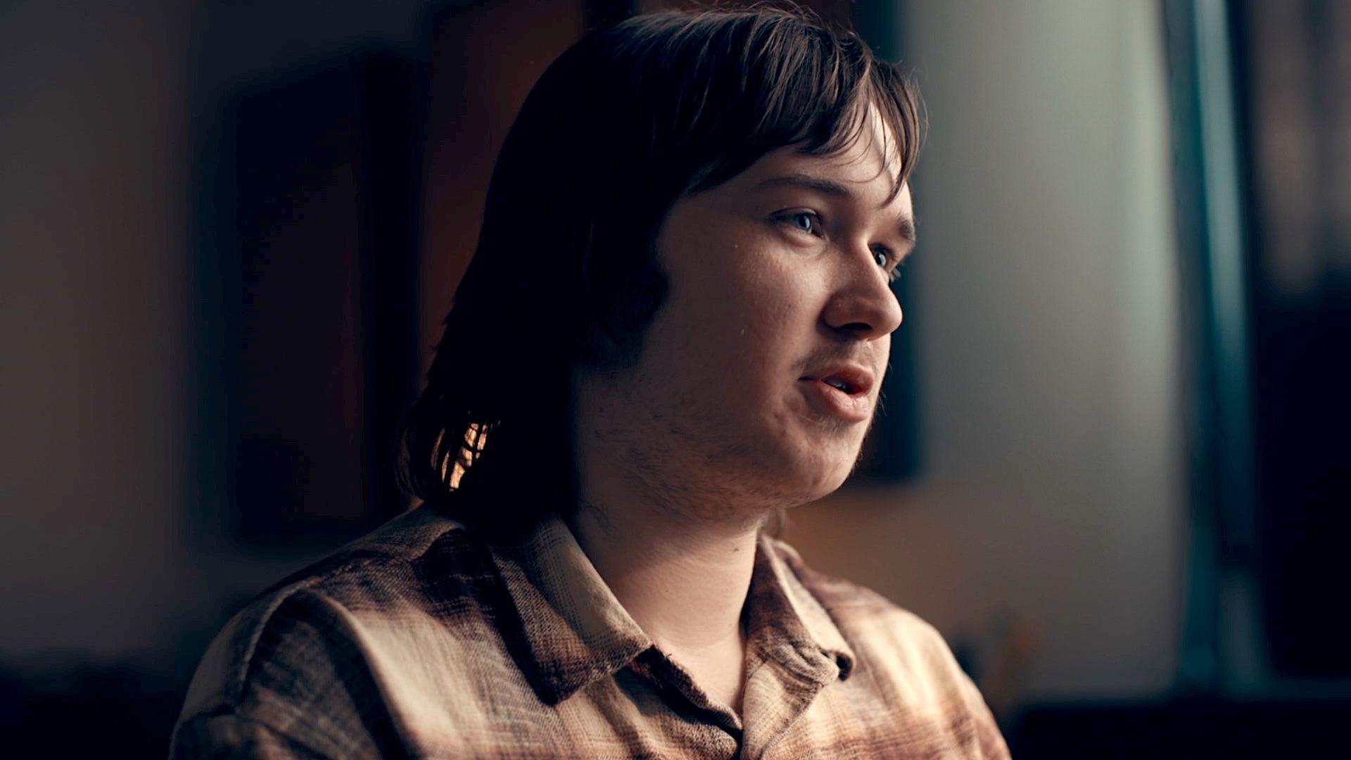 Calum, a young man with long brown hair and slight facial hair, wearing a brown checked shirt, pictured in close-up looking to the right of the camera in a darkened room with the curtains behind him drawn, stark light falling across his face from the right of the image.