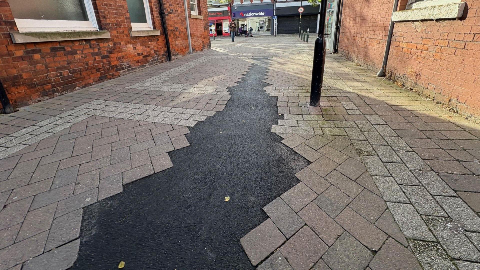 A pavement with a patch of dark asphalt running through the middle of paving stones. On both sides are red brick buildings. At the end of the path, in the distance, are three commercial premises, including a Nationwide building society.