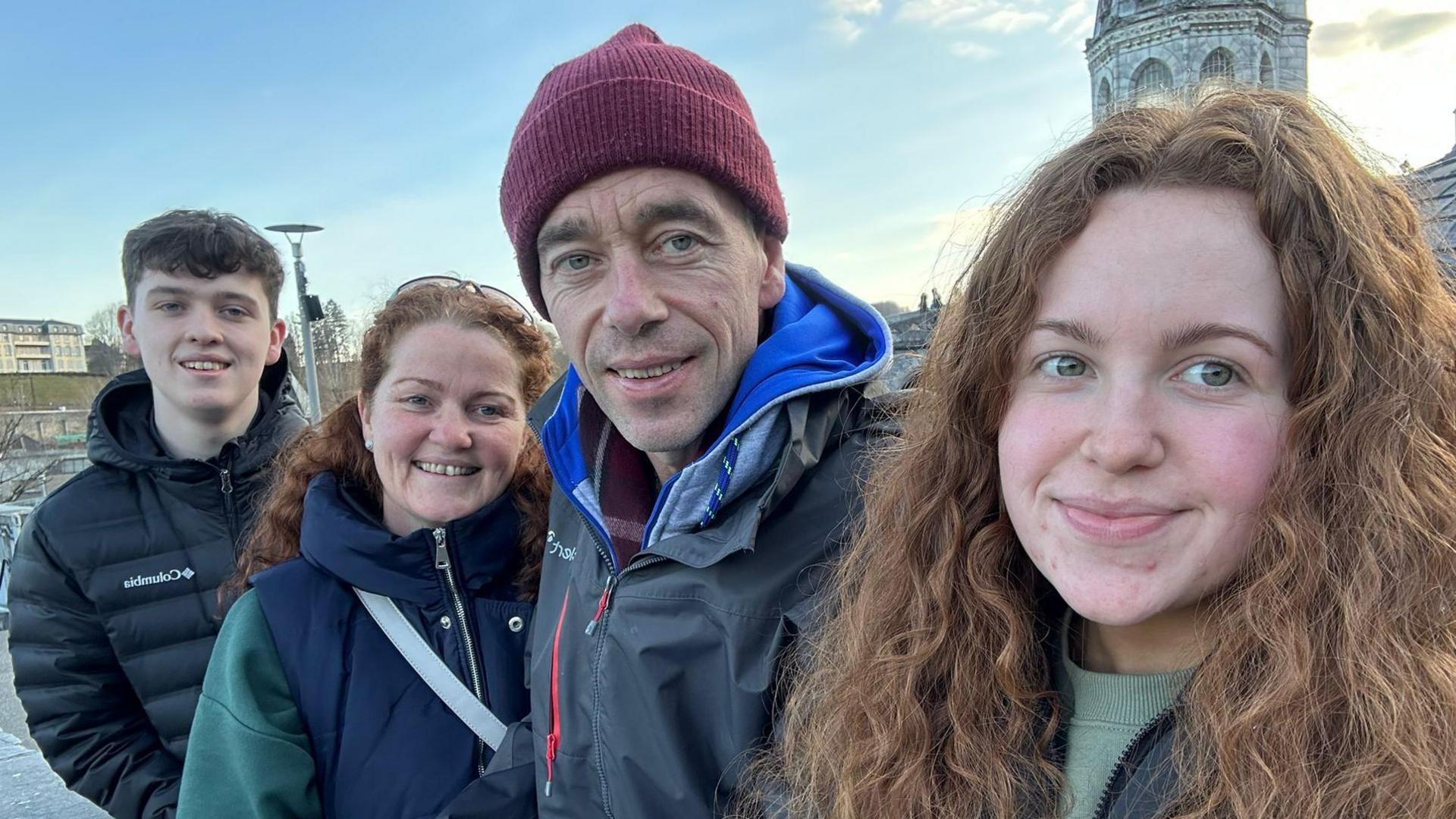 (L-R) Tiarnán, Debbie, John and Erin Gallagher are standing side by side. 
Tiarnán has black hair and is wearing a black hooded jacket.
Debbie us wearing a navy and green jacket.
John is wearing a red hat and blue hooded jacket, while Erin, who has long curly brown hair, is wearing a green jumper and jacket. 