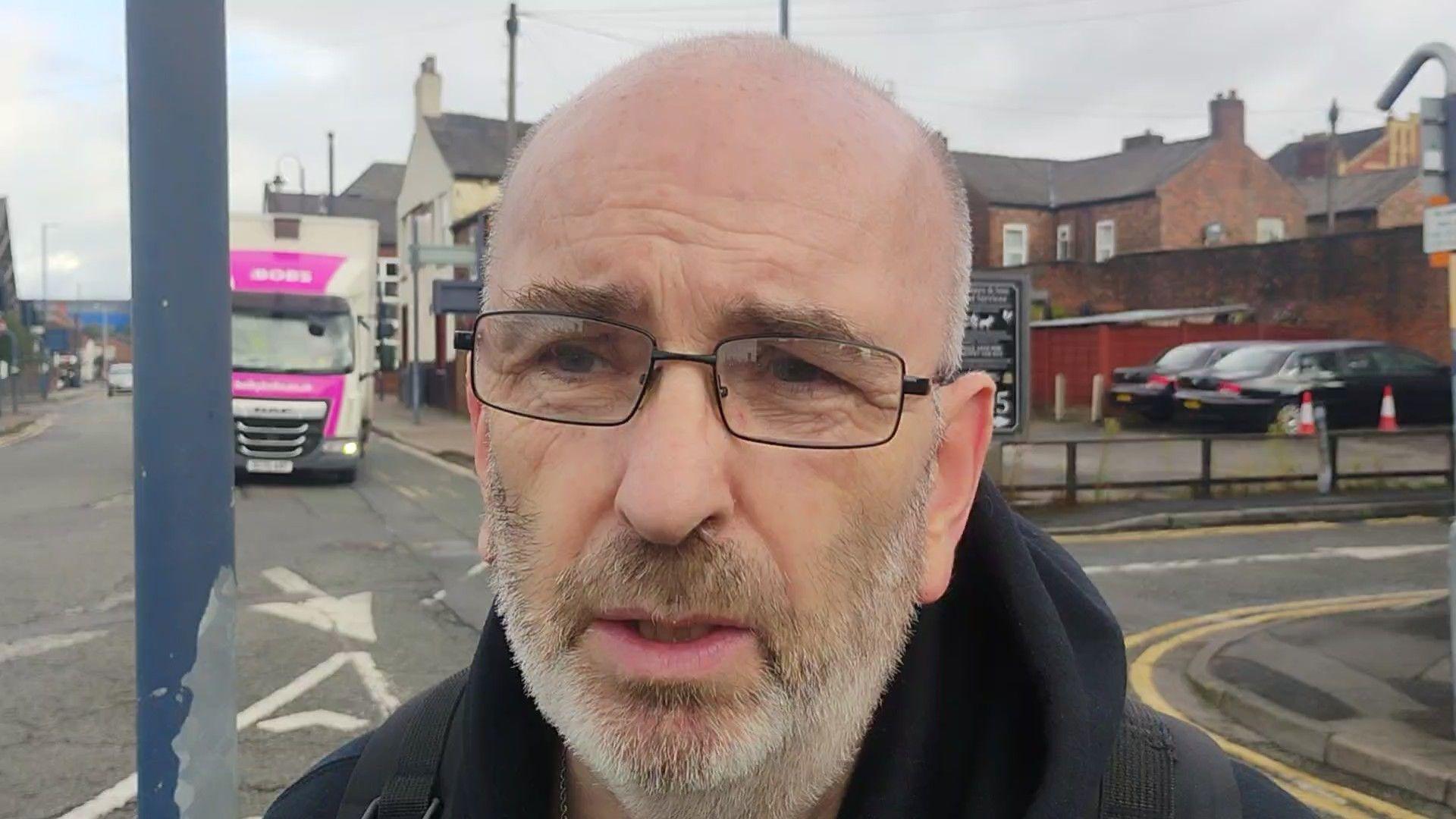 Peter Smith, who has a bald head, black-framed rectangular glasses, and a short white beard, speaks to the camera on a busy road while wearing a black hoodie.