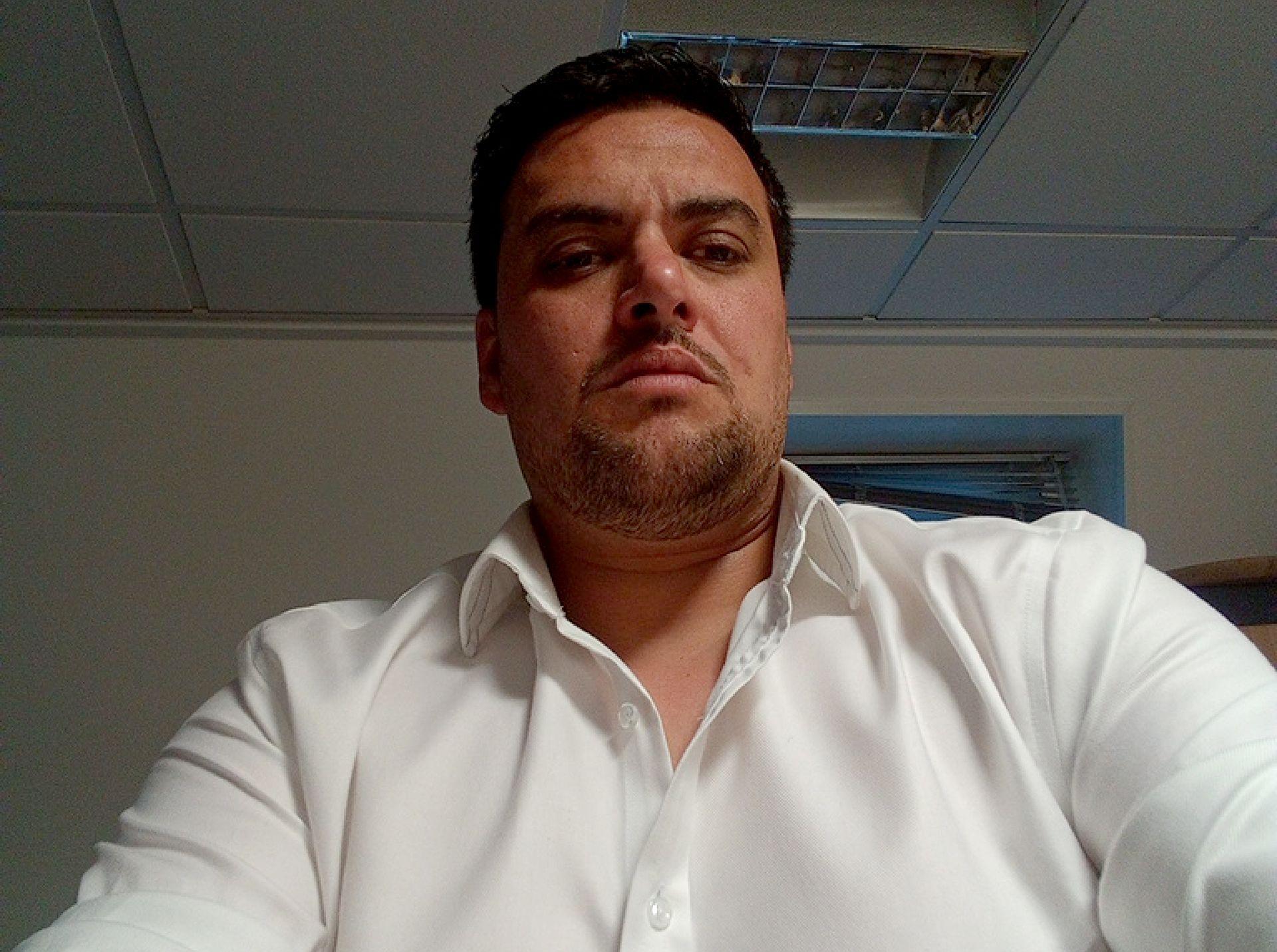 Dominic D'Sa: A man with dark hair and stubbled wearing an open-necked white shirt