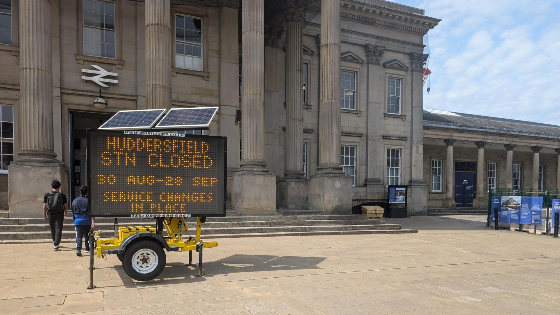 The exterior of a railway station during the day with a digital billboard outside which says 'Huddersfield stn closed 30 Aug-28 Sept service changes in place'.