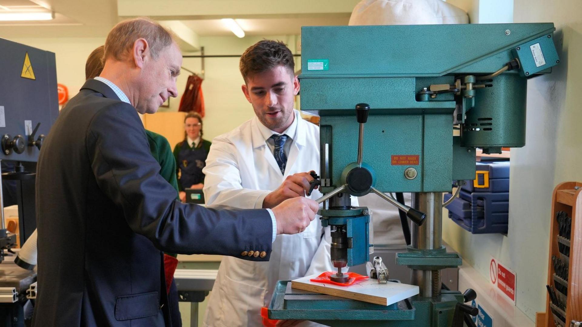 The Duke of Edinburgh, Prince Edward, helps put a Joint Award Initiative logo on a plastic orange bowl. He is in a Technology and Design classroom and is joined by the teacher of the class. 