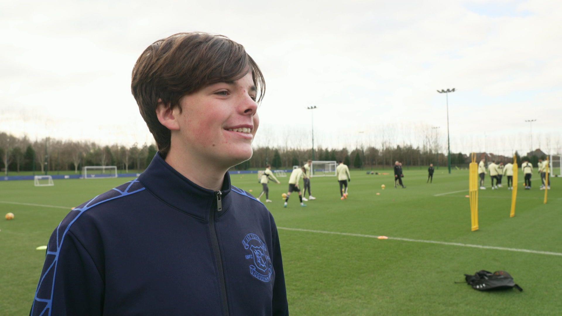 Mackenzie, who has brown hair and wears a zip up Everton branded jacket, smiles at the camera as Everton players train on a grass pitch behind him