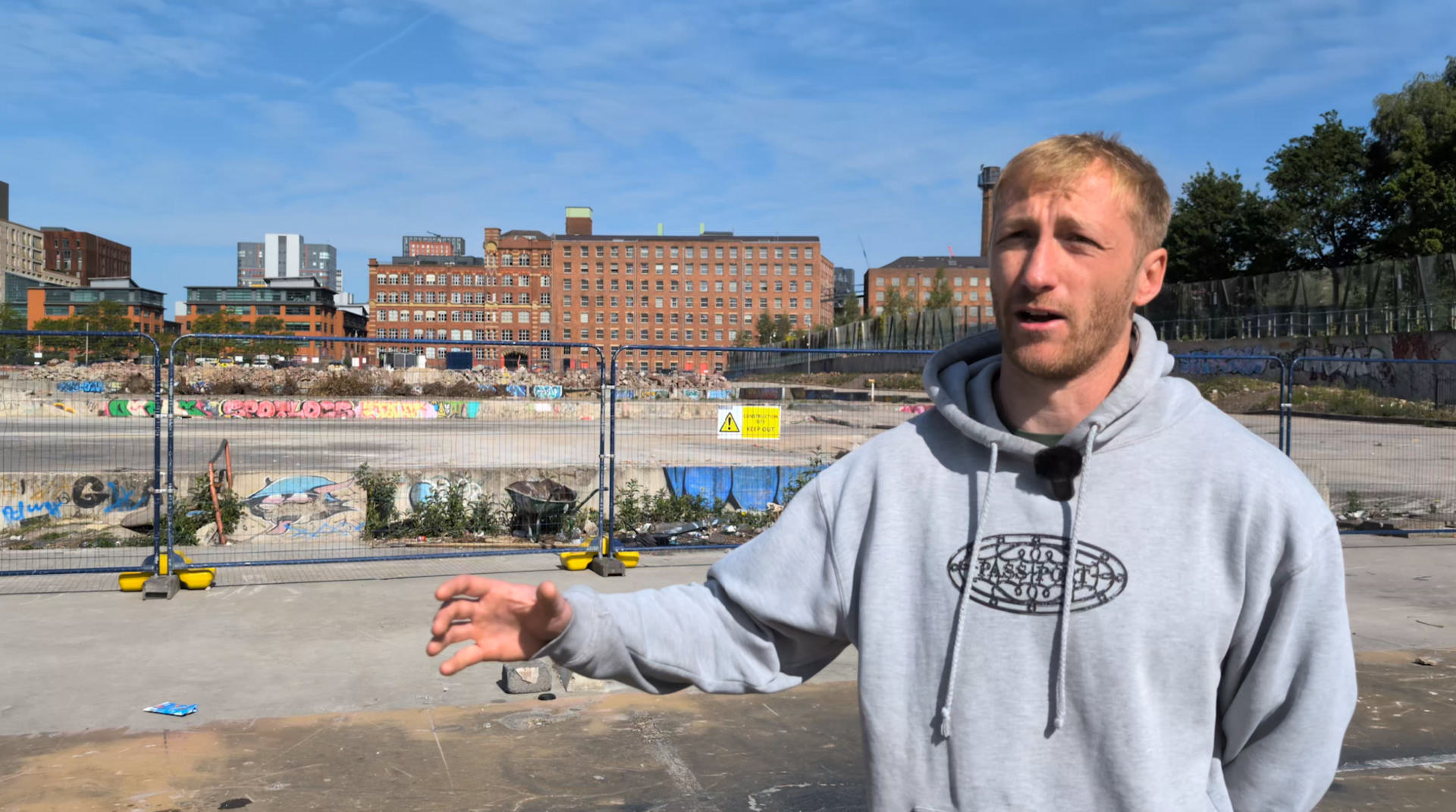 A man in a grey hoodie with his right arm outstretched stands in front of the demolished skatepark, which is fenced off.