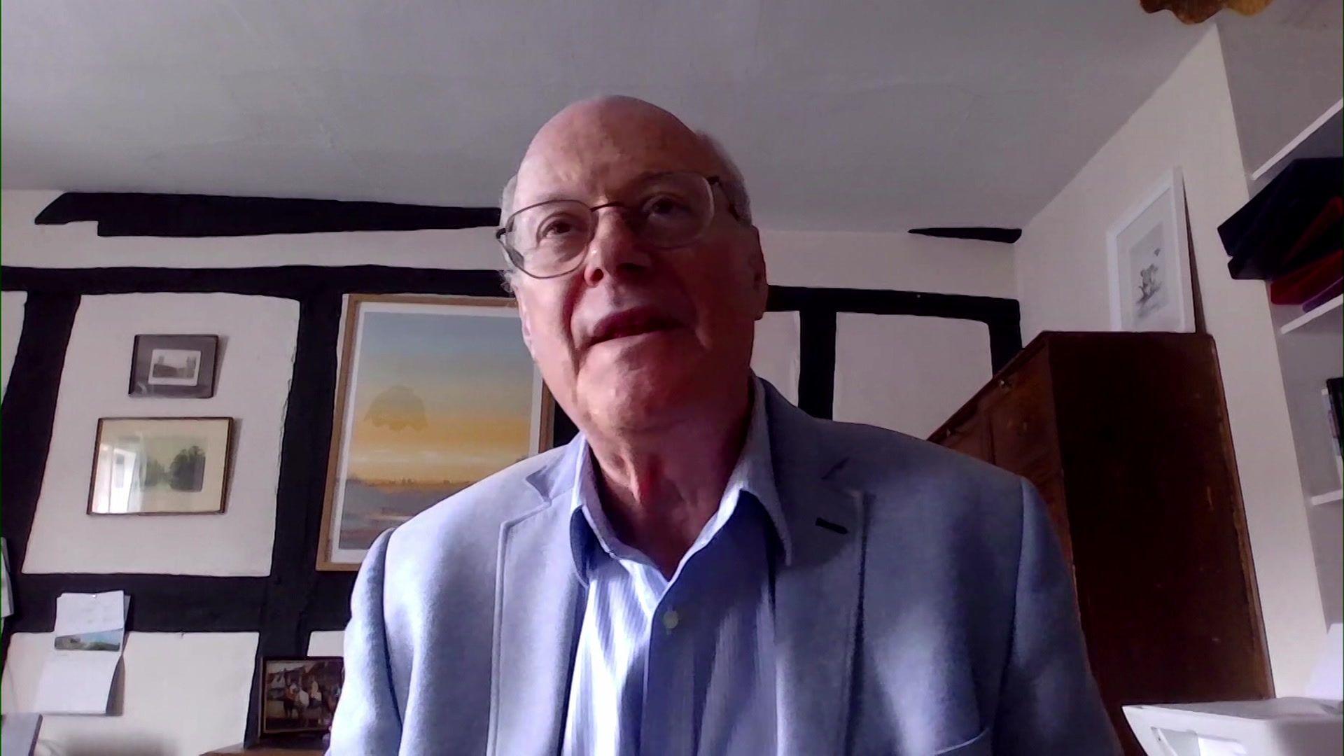 Sir John Saunders, sitting in what appears to be an office in a Tudor-style cottage, is wearing a light blue-violet jacket and speaking into the camera on a Zoom meeting