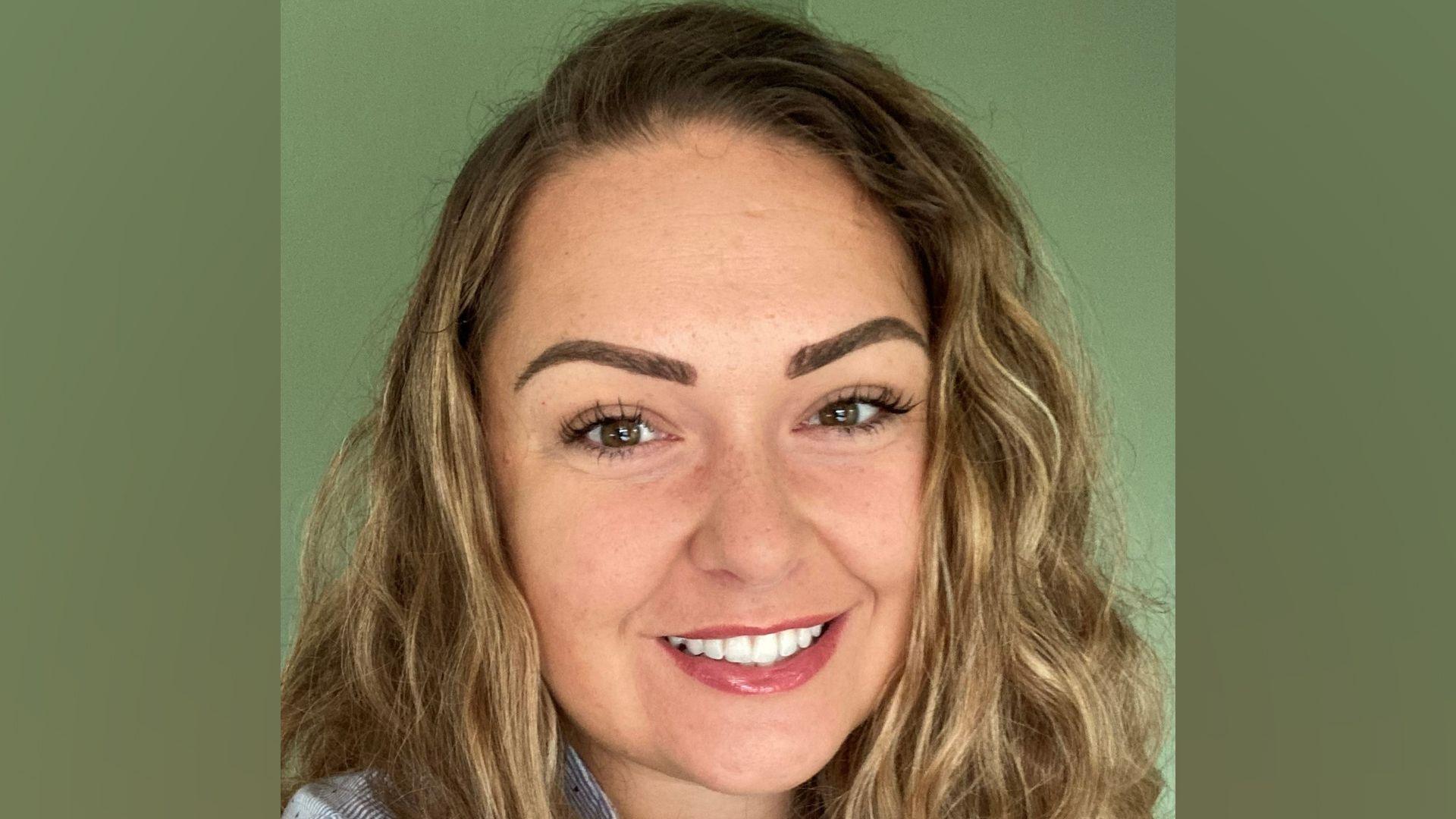 A head shot of a woman with wavy fair hair, smiling and looking into the camera. She is in front of a green background.