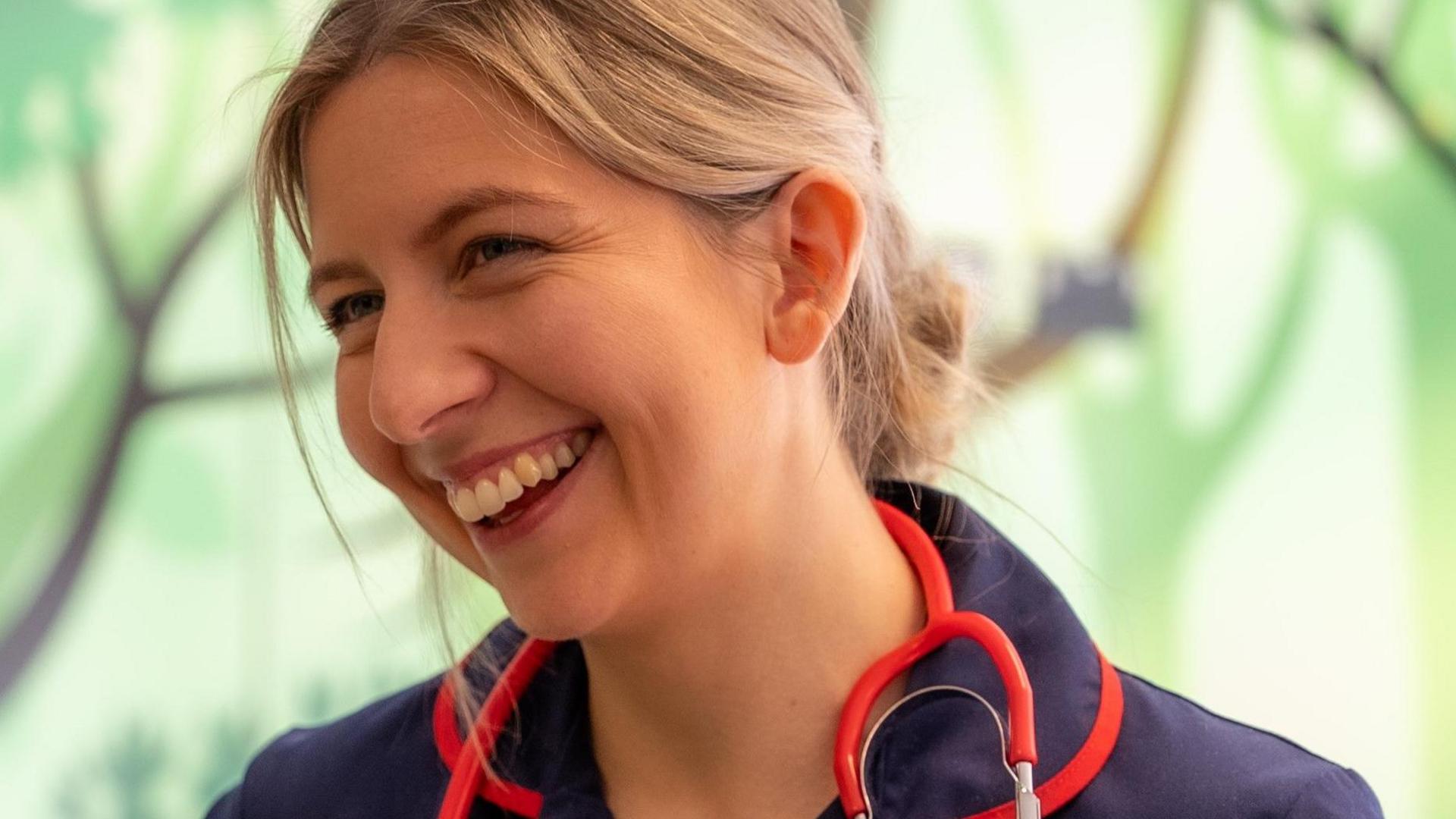 A woman with blonde hair that is tied back into a bun smiles away from the camera. She is wearing a navy nurse uniform with a red stethoscope around her neck. 