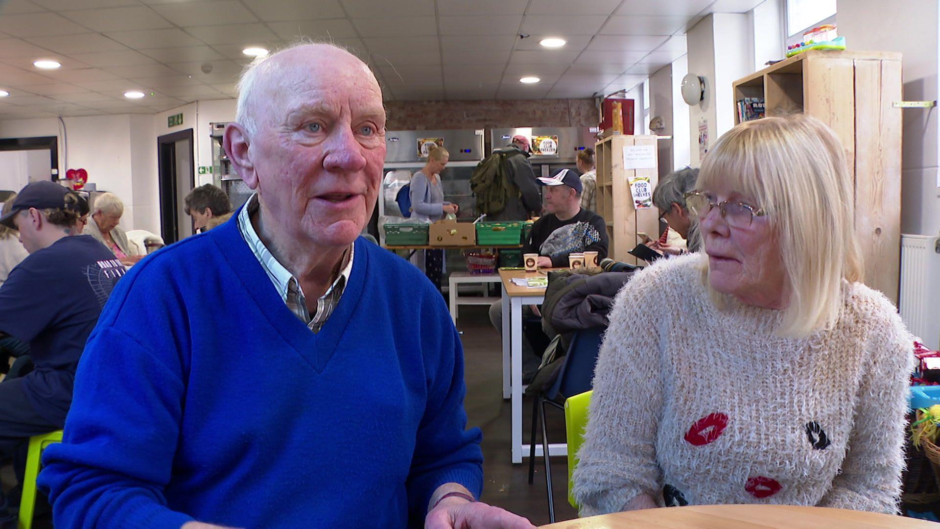 Man in blue jumper with grey hair sat at table next to woman in grey jumper with blonde hair and glasses.