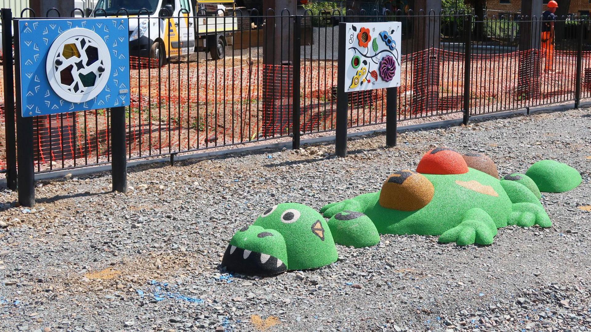 A play area, showing a green dinosaur and play equipment, with grey slate concrete ground. Building work is going on behind the play equipment.