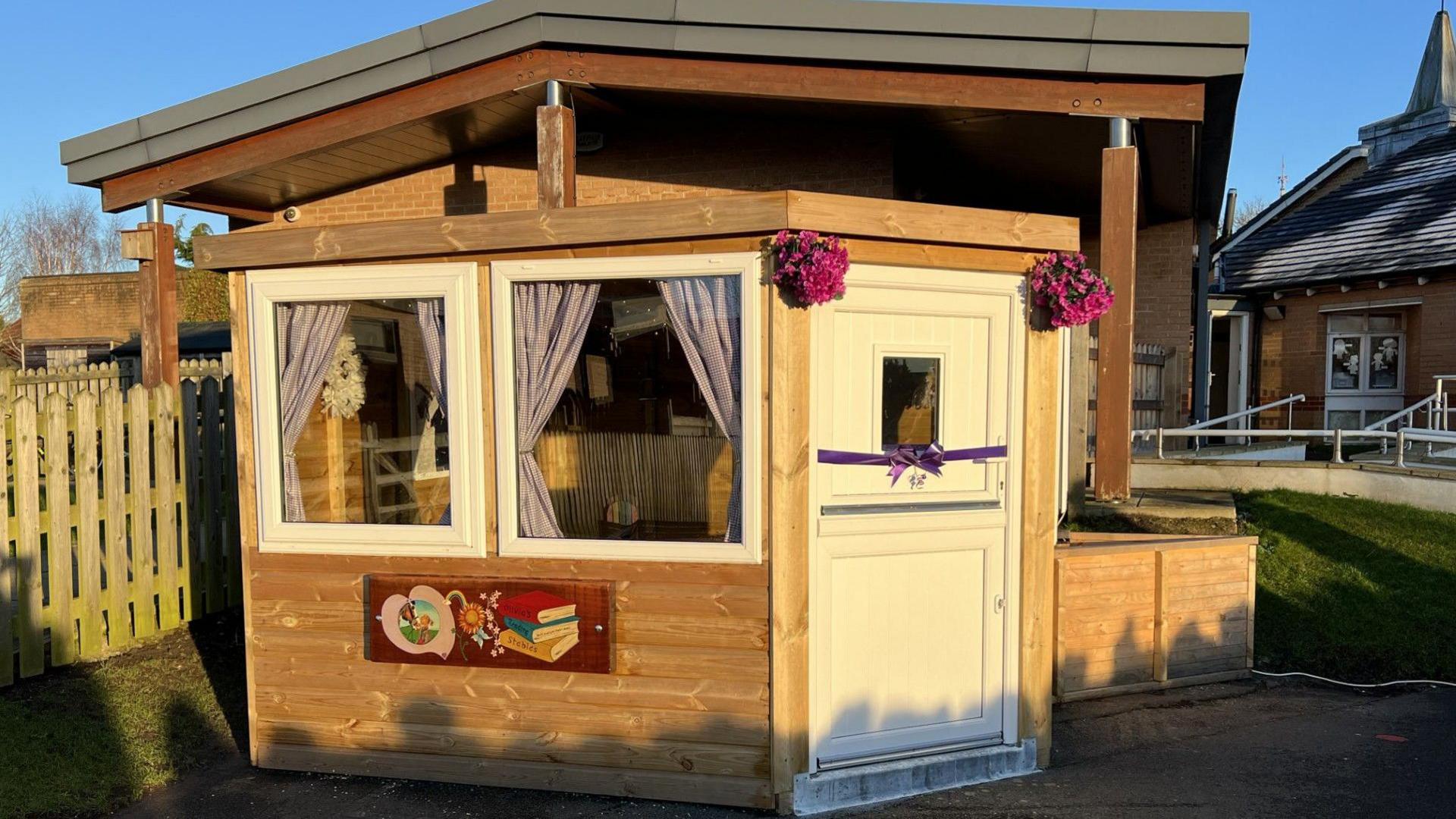 A wooden hut building with two white square windows to the right of the front of building and a white stable looking door to the right with two round bunches of purple flowers above the door. Under the two windows is a wooden plaque showing a picture of Olivia with her horse in a pink heart with a painting of three books next to her.  