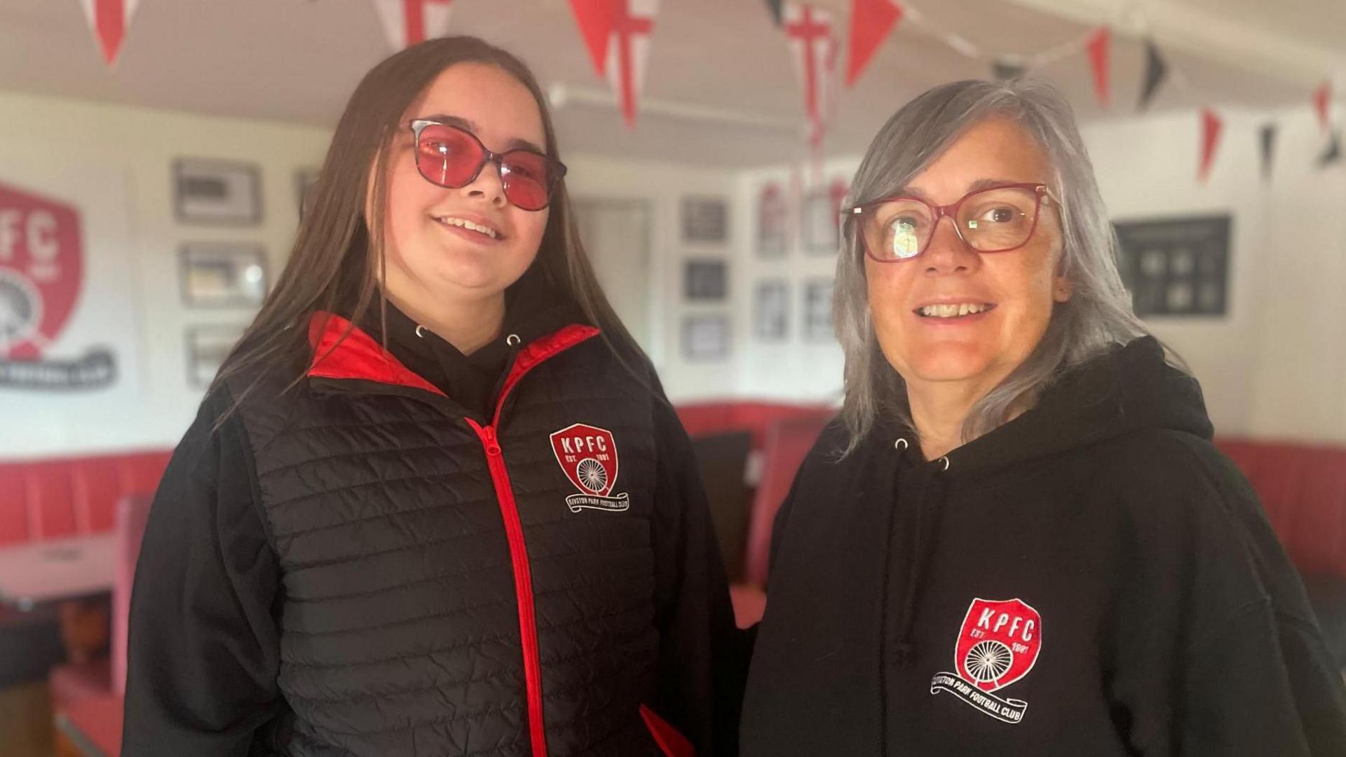 A young woman in red glasses and an older woman stand together in matching black and red sports coats