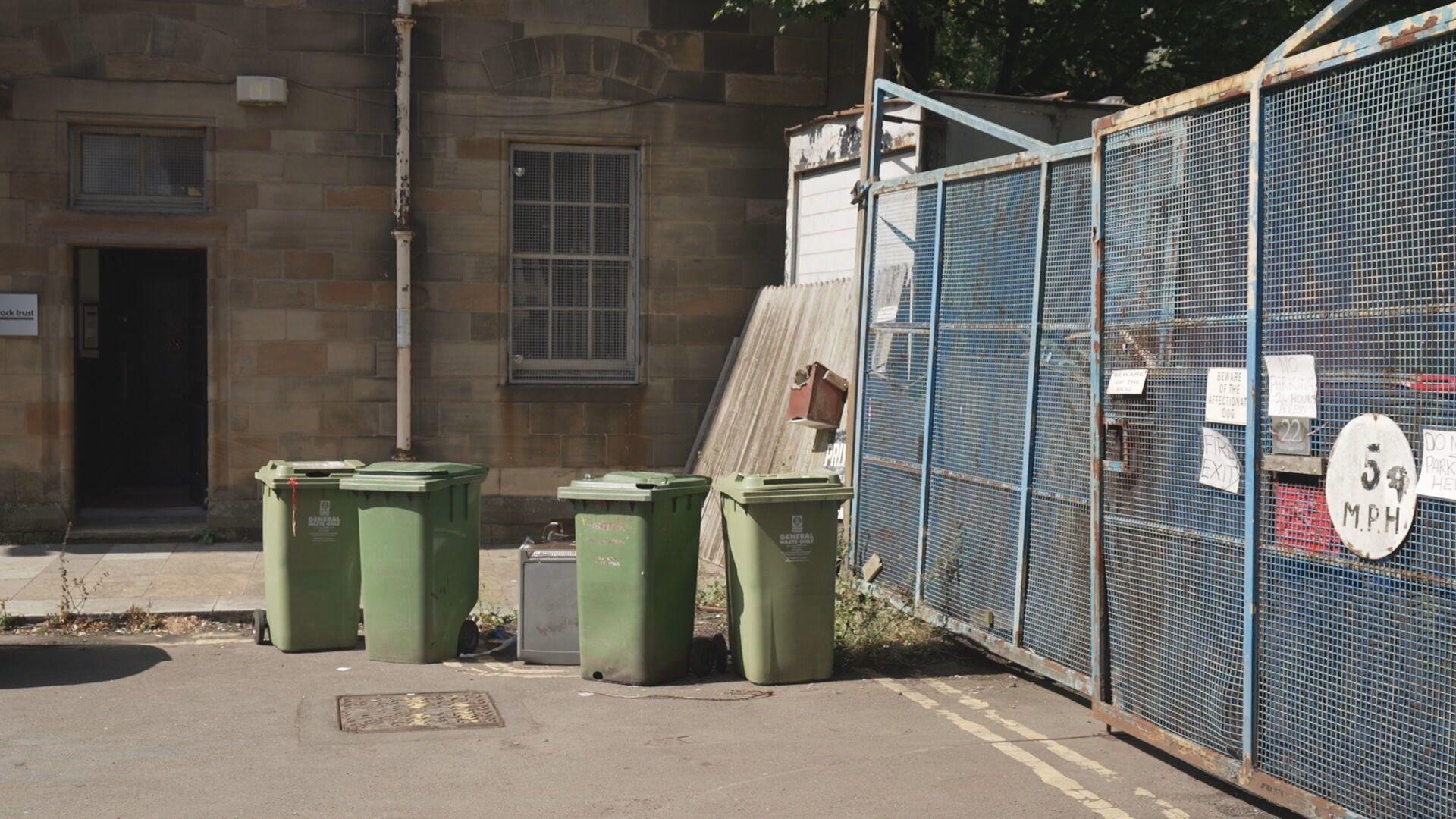 The entrance to the Stringfellows' yard is a locked metal gate, with warning signs about a dog and 5mph speed suggestion. Four green wheelie bins sit in a haphazard fashion outside the gates. Tenement flats run up to the entrance of the yard.