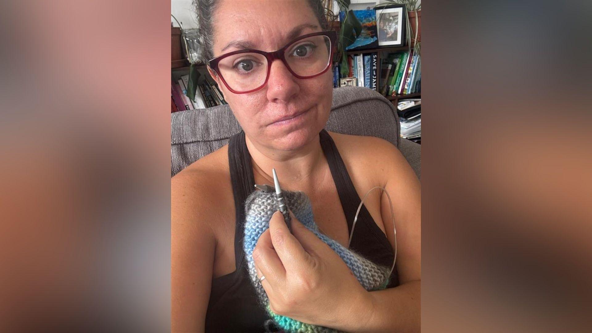 Mairead is wearing glasses and knitting. She is sitting on a sofa with a bookcase behind her.