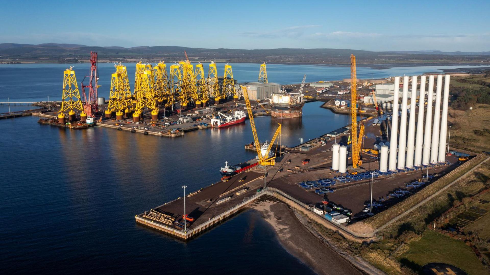 Bright yellow jackets for offshore wind turbines and white turbine towers are stacked on parts of the yard. The sea around the port is flat calm and the sky above is blue.