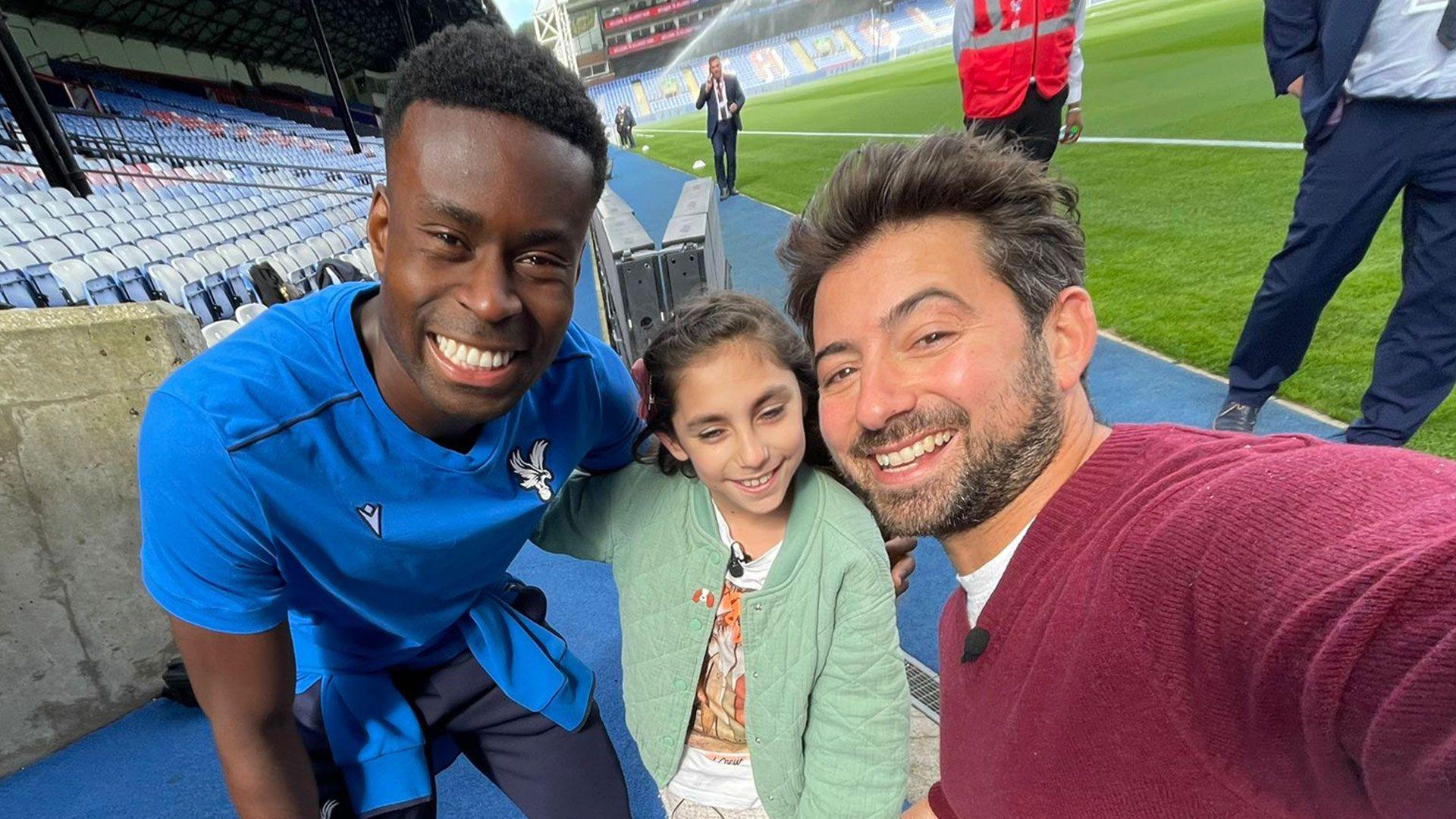 Crystal Palace captain Marc Guehi next to young fan Sophia and Newsround presenter Ricky, smiling for the camera