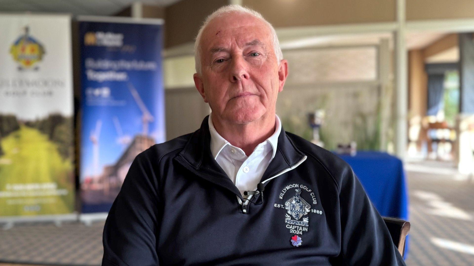 Killymoon Golf Club Captain Anthony McCann sitting down. He is wearing a navy blue golf club sweatshirt and white short. He has grey hair.