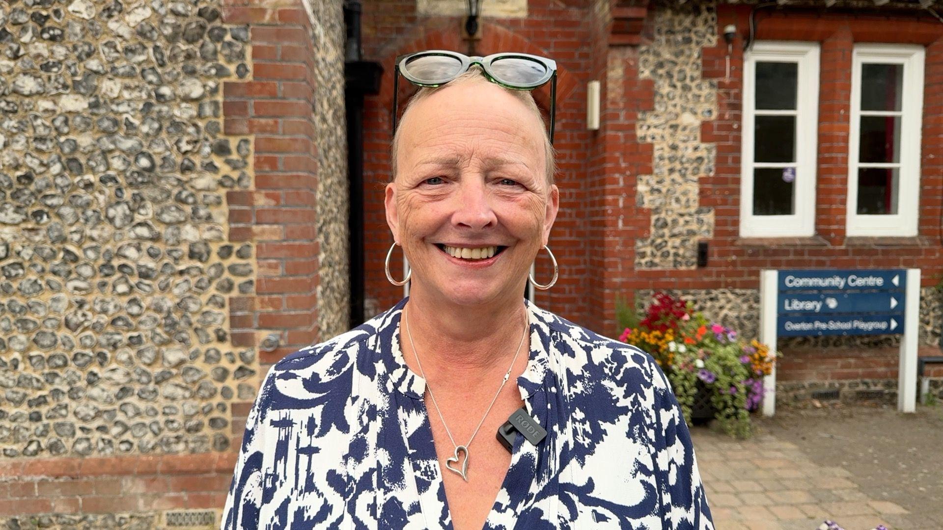 A woman with very short, grey hair, sunglasses on top of her head, gold hoop earrings, and wearing a white and blue patterned blouse, standing in front of a council building and smiling.