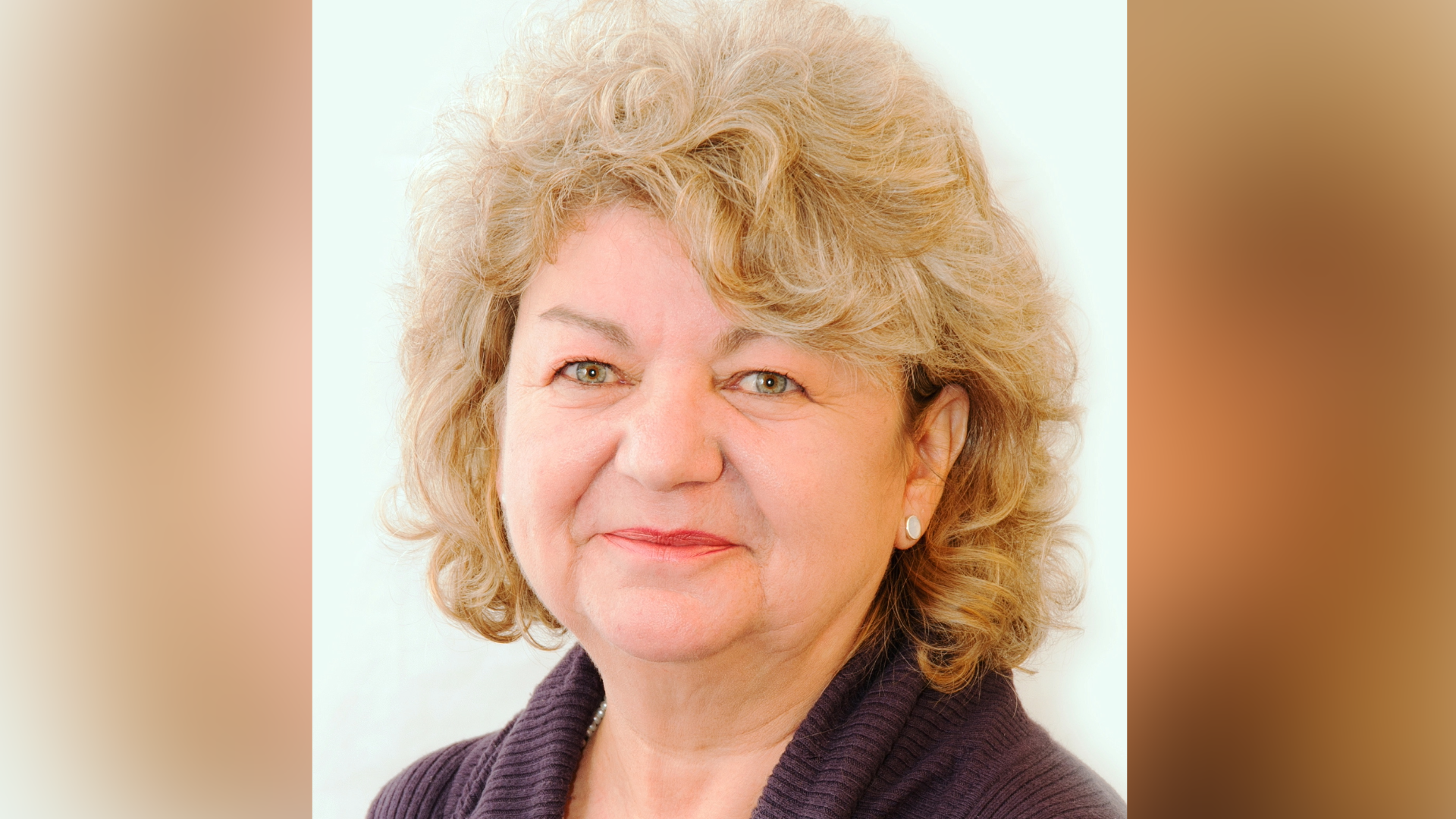 A woman with short curly blonde hair is pictured smiling while wearing a brown cardigan. 