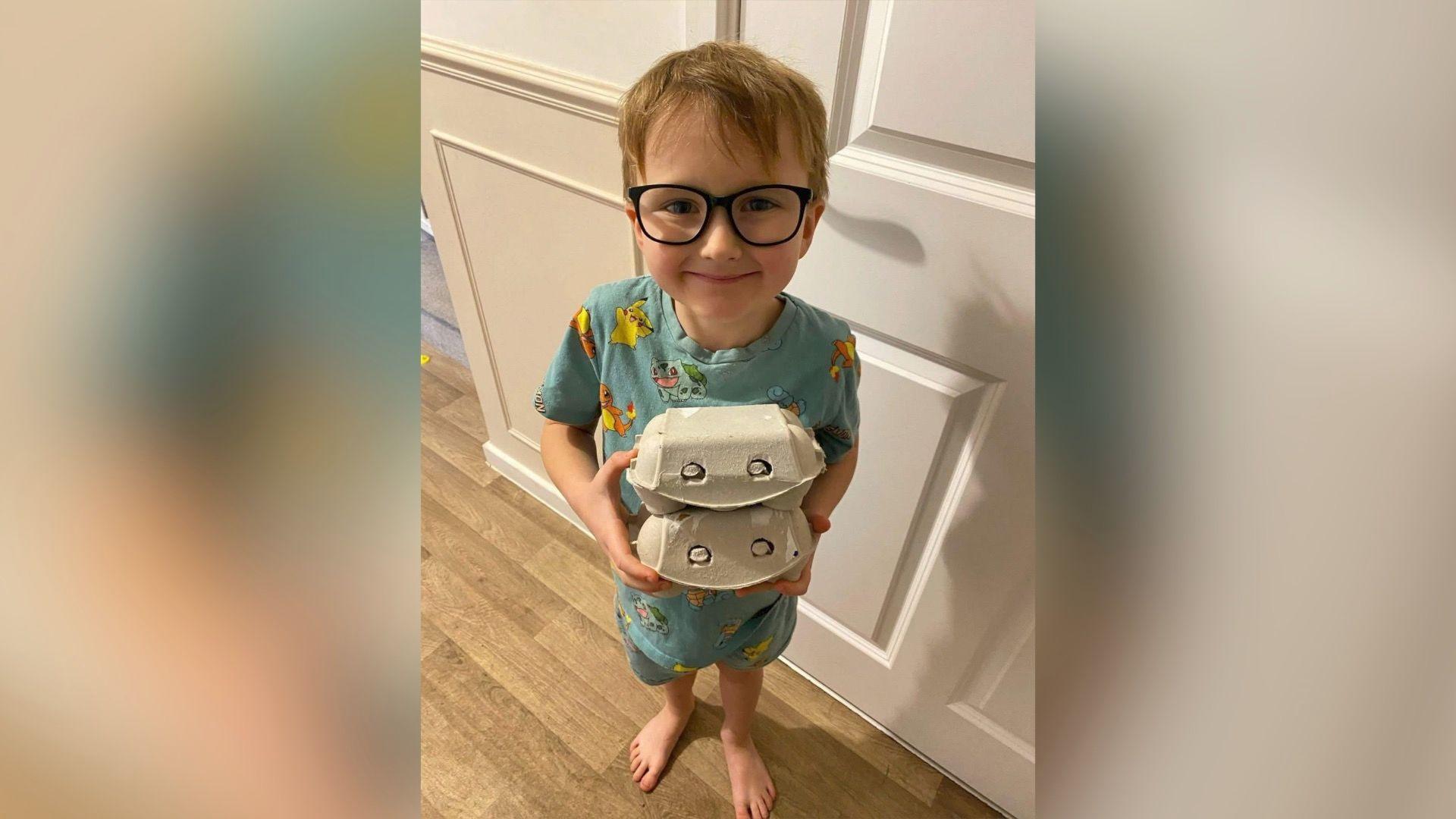 A boy smiling straight at the camera while holding two egg boxes. He is wearing blue pyjamas and glasses.