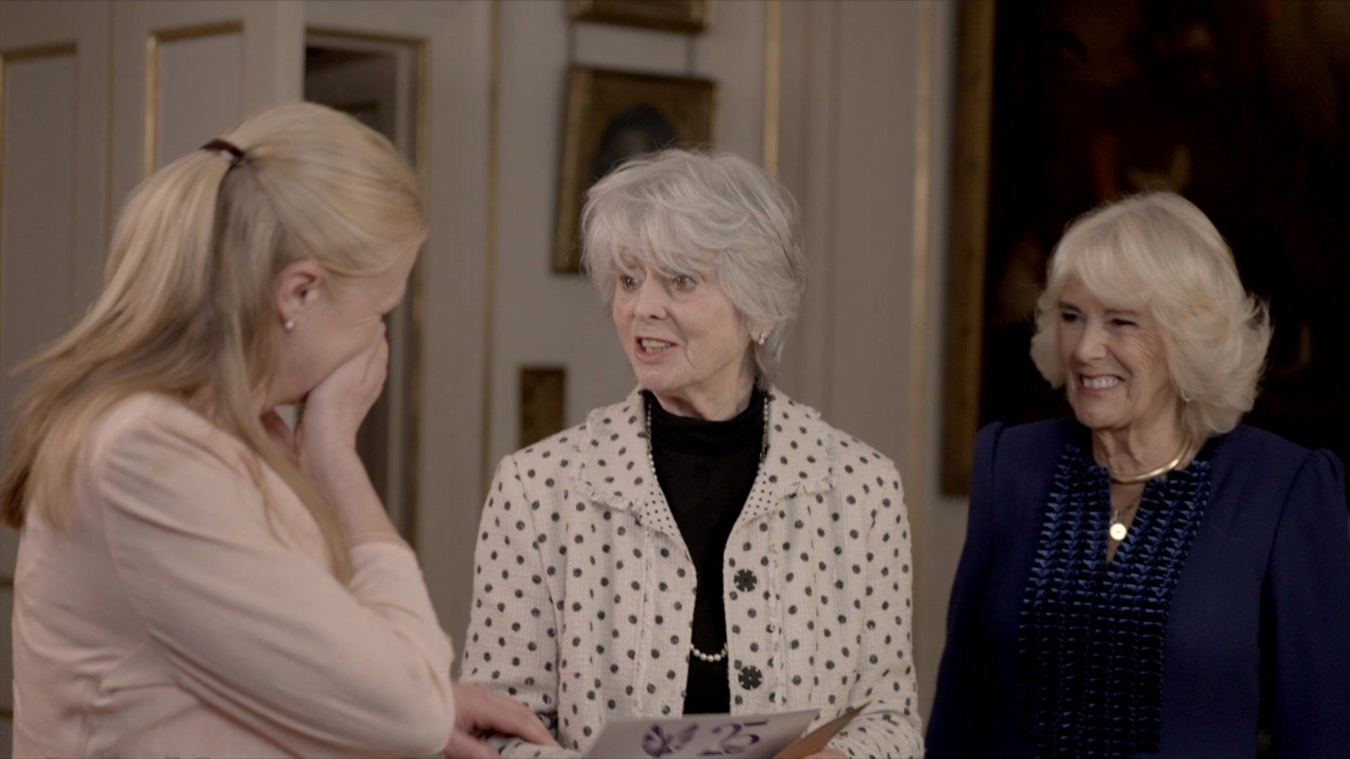 Hetti Barkworth-Nanton, who is not facing the camera, has her blonde hair tied back and has her hand to her face in surprise. She is wearing a pale pink jacket. Diana Parkes is looking at her in surprise, having opened a card informing them that they have been given a Pride in Britain award. She is wearing a white jacket with blue dots. The Queen is to Ms Parkes' left and is smiling. She is wearing a dark blue jacket. 