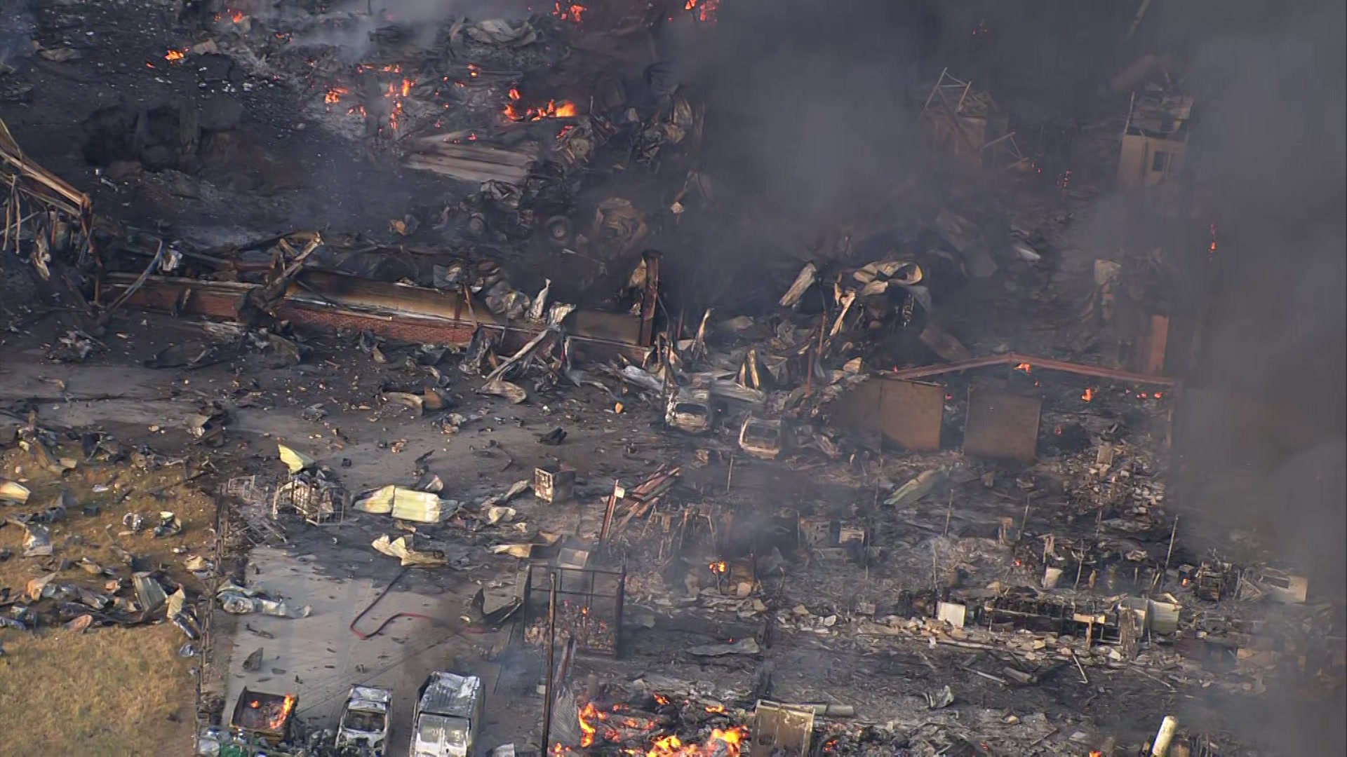 An aerial view of an industrial sit covered in debris. There are flames dotted throughout the site and black smoke rising.