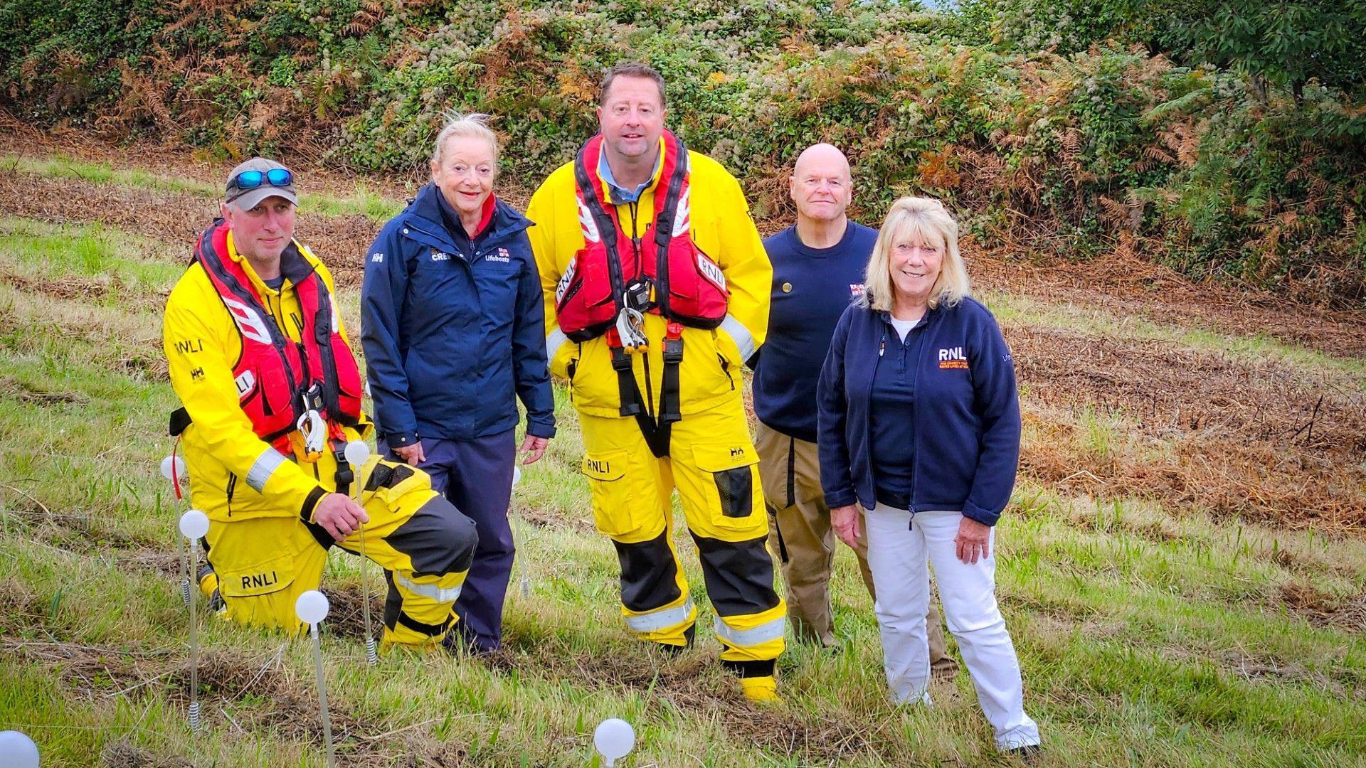 A team of five RNLI volunteers stand on a grassy bank. They are all smiling at the camera. In front of them are some of the fibre optic stems they've been planting.