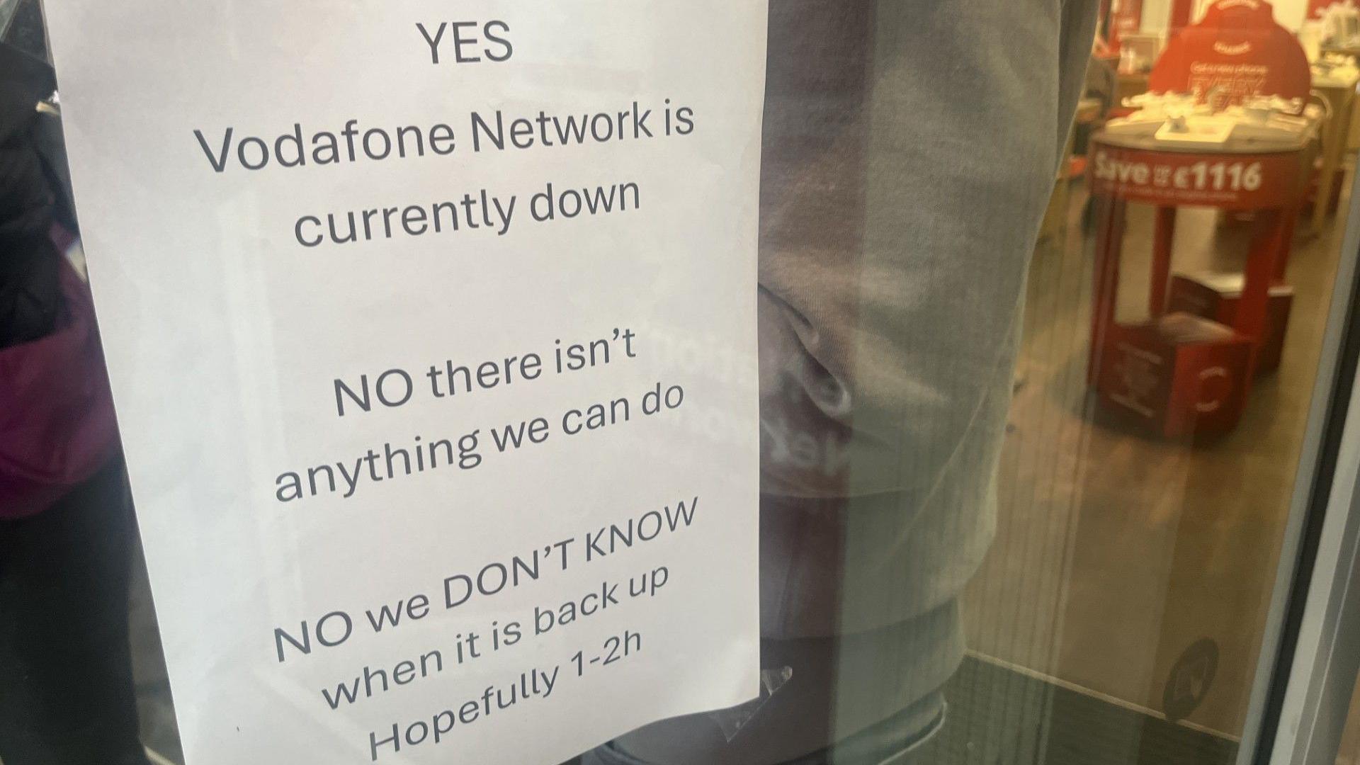A piece of A4 paper stuck to the inside of a window of a Vodafone shop states: "YES Vodafone Network is currently down. NO there isn't anything we can do. NO we DON'T KNOW when it is back up. Hopefully 1-2h"
