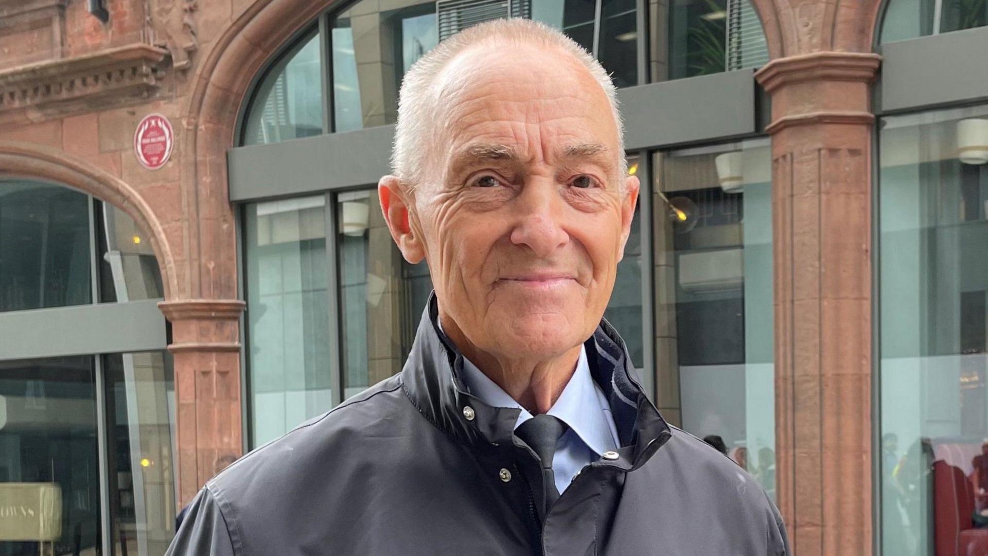 Graham Brown, a man with short hair and dark eyes wearing a blue shirt and tie and a jacket. He is stood in front of a building and the red plaque can be seen on the wall behind him.