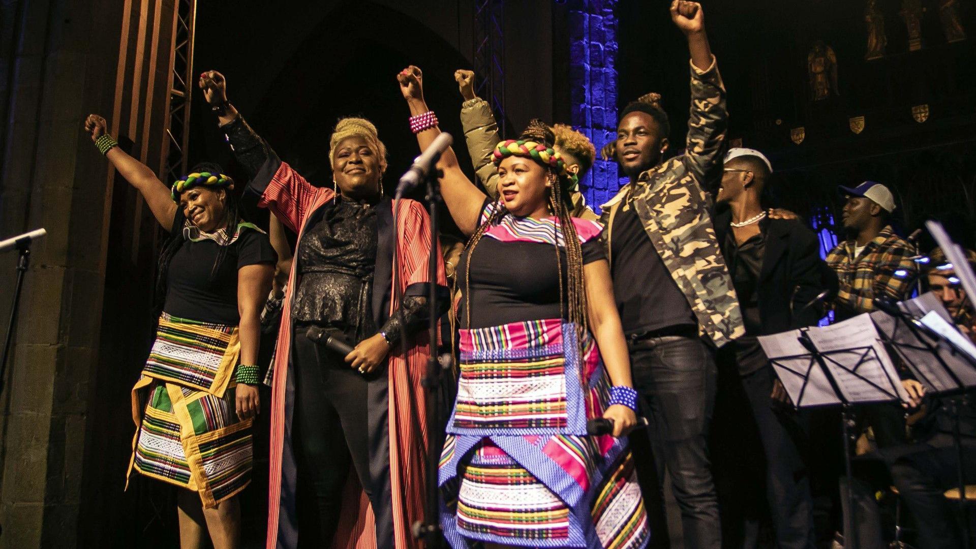 Soweto Gospel Choir, Voices of Virtue and Kema Kay on a stage. Members of the group are holding their fist in the air.