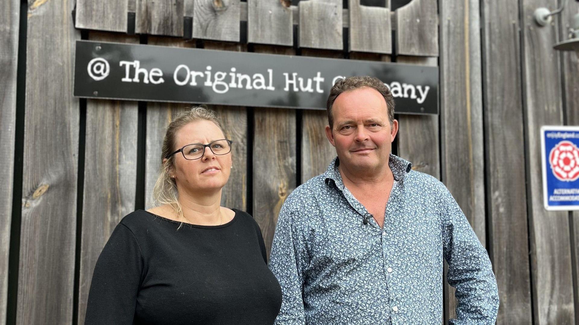 A man and woman stand outside a cafe. She has blonde hair in a ponytail and is wearing a black top and black glasses. He has brown hair with a blue floral shirt on. There is fencing behind them, with a sign saying The Original Hut Company