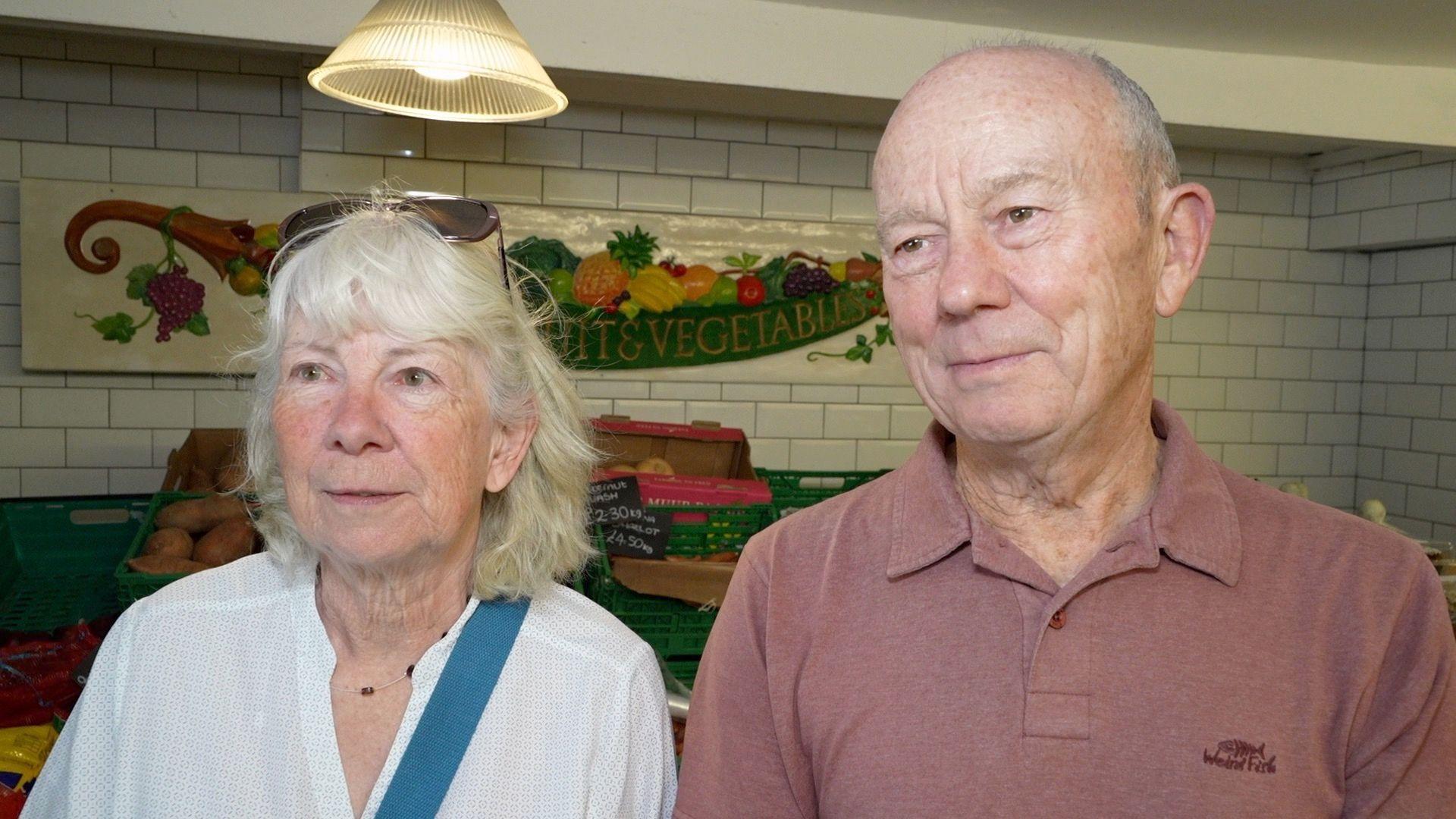 Helen and Robert Flindall smile as they look slightly away from the camera. Mrs Flindall has shoulder length grey hair and wears a white blouse with sunglasses on her head, a necklace around her neck and a blue bag strap around her body. Mr Flindall is bald and wears a red polo T-shirt.