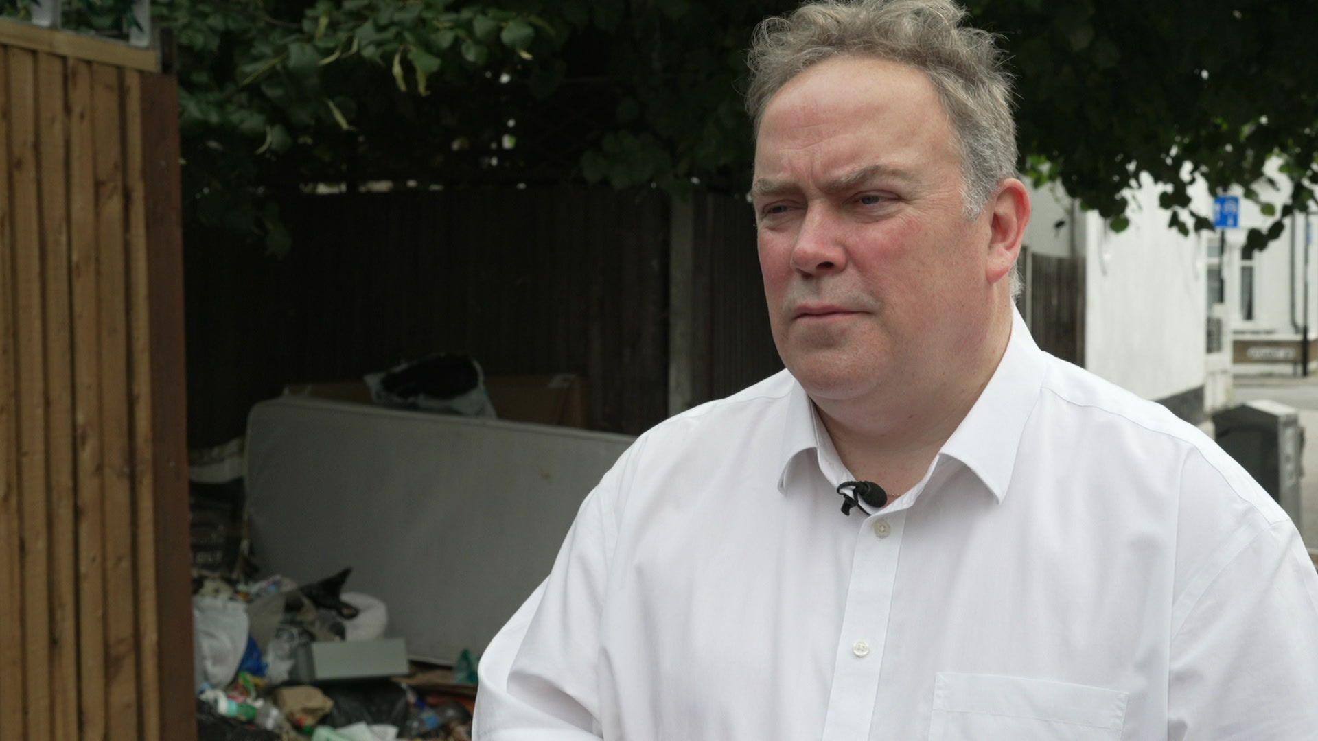 Jason Perry, who has grey hair and is wearing a white shirt, stands next to a pile of rubbish on a street