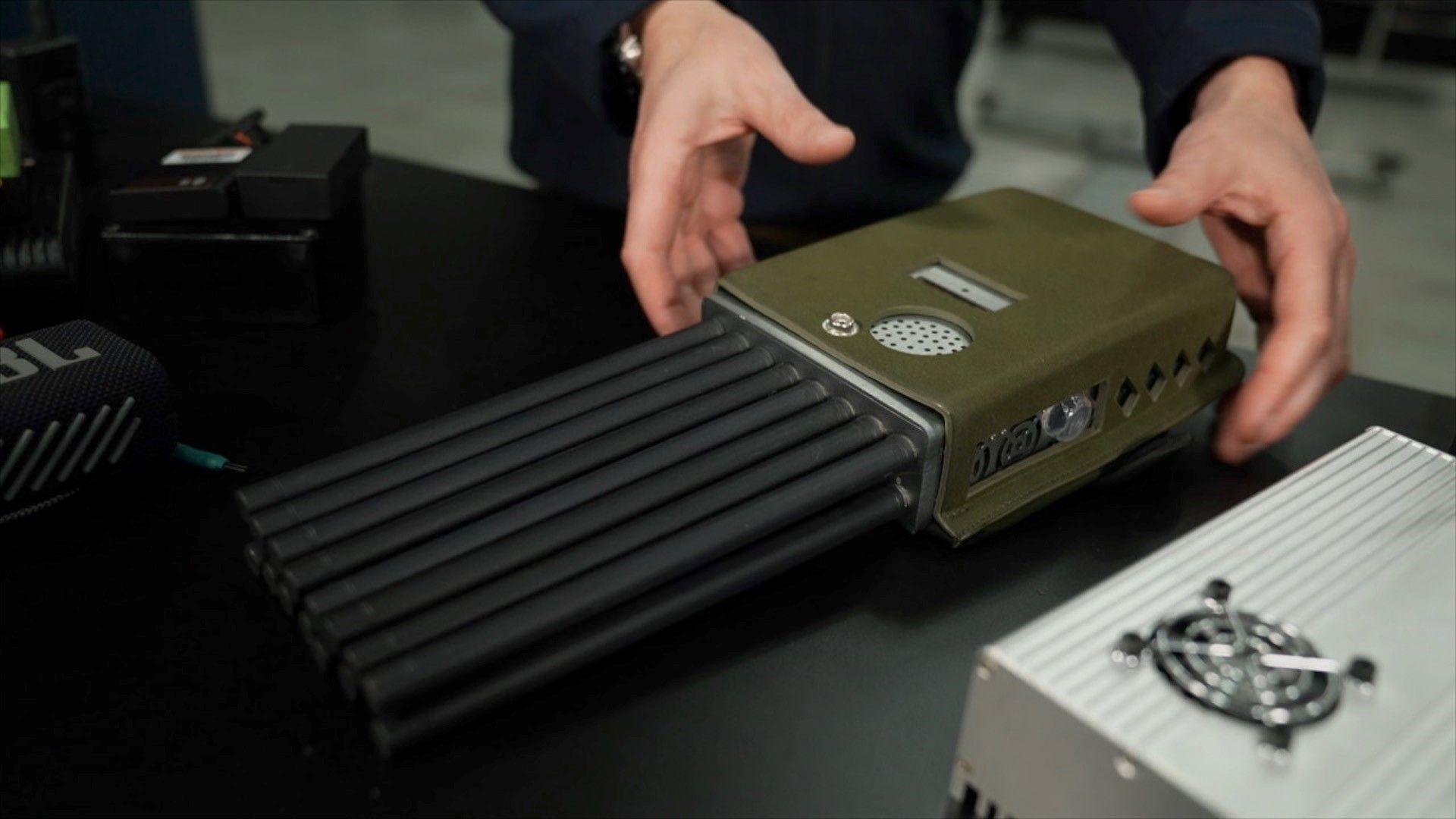A person’s hands are positioned near a rectangular electronic device with an olive-green casing and multiple black cylindrical antennas extending from one end. The device has a small screen, a speaker grille, and several ports on its surface. It is placed on a black table alongside other electronic equipment.