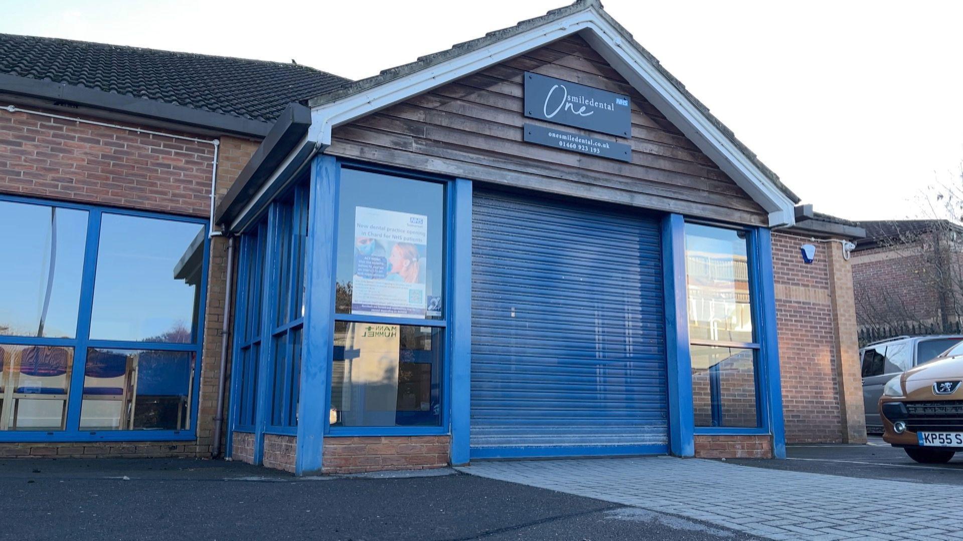The exterior of a the new dentist practice. It has blue shutters and a black sign saying 'One Smile Dental'.