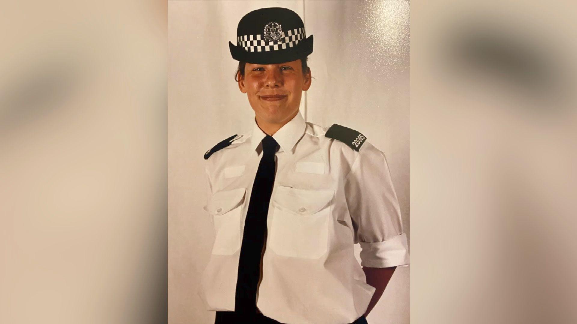 Kerry Snuggs poses for a photo wearing her police uniform. She is smiling at the camera and stands, with her hands behind her back. She wears a black bowler style hat with a black check ribbon around it and the police logo on the front, a white collared shirt with black pads on the shoulders and a black tie.