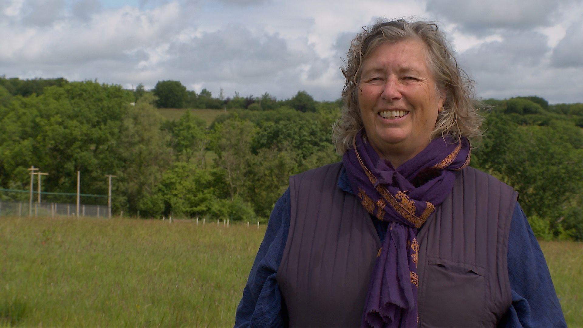 Dorette Engi, smiling and wearing a blue jumper with purple scarf and gillet. She is standing in her land with the stork avery in the background.