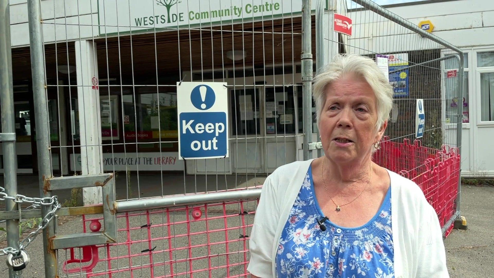 Nicky Skeat stands in front of the community centre, which is fronted by a padlocked metal fence and a sign saying 'keep out'. She has short, white hair and wears a blue blouse and a white cardigan.