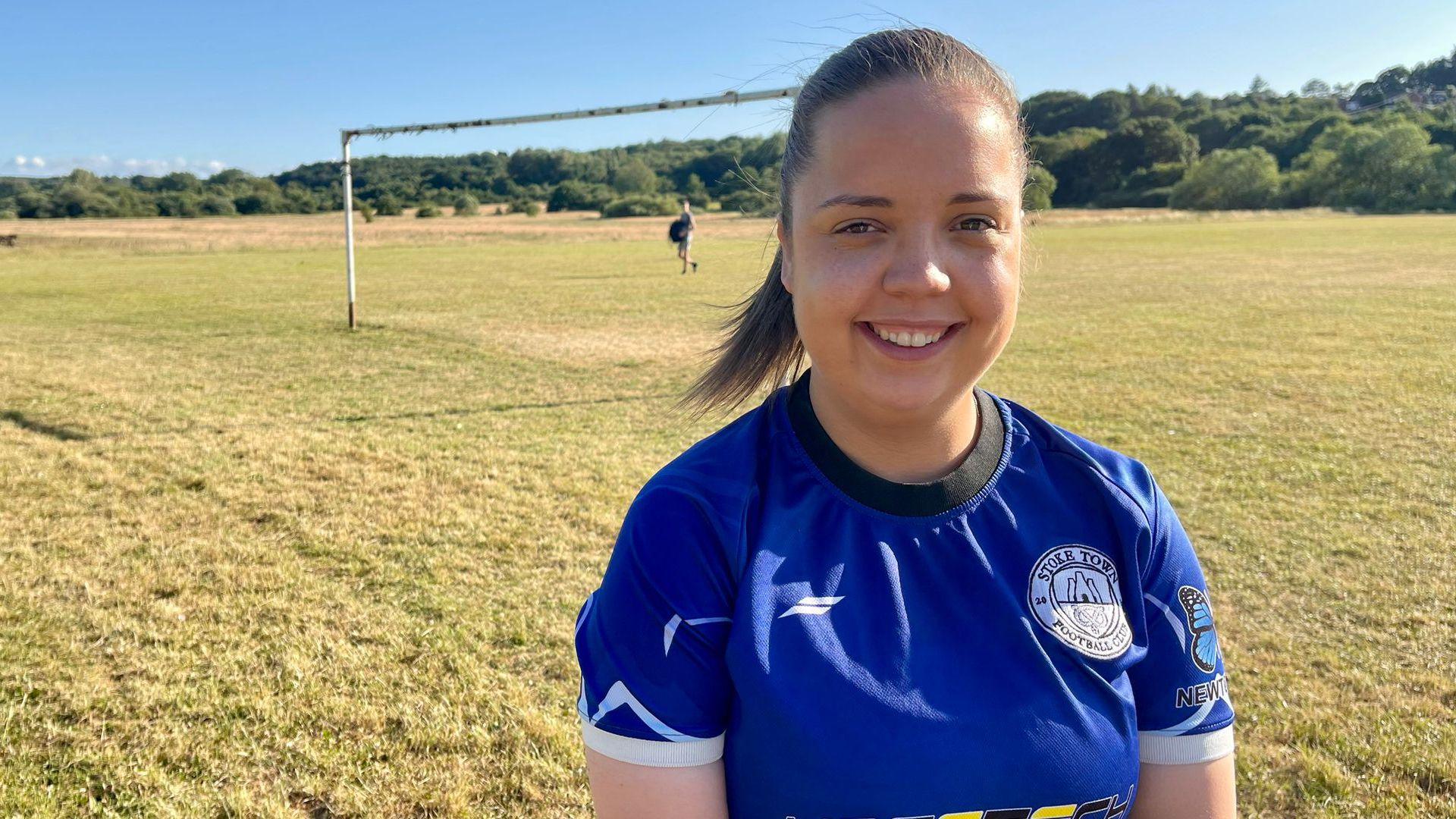 A woman in blue football shirt stands on a grass roots football pitch. She has dark hair tied in a ponytail. Her shirt displays the club badge of Stoke Town Football Club and a clothing logo. Rusty goal posts and trees can be seen in the distance.